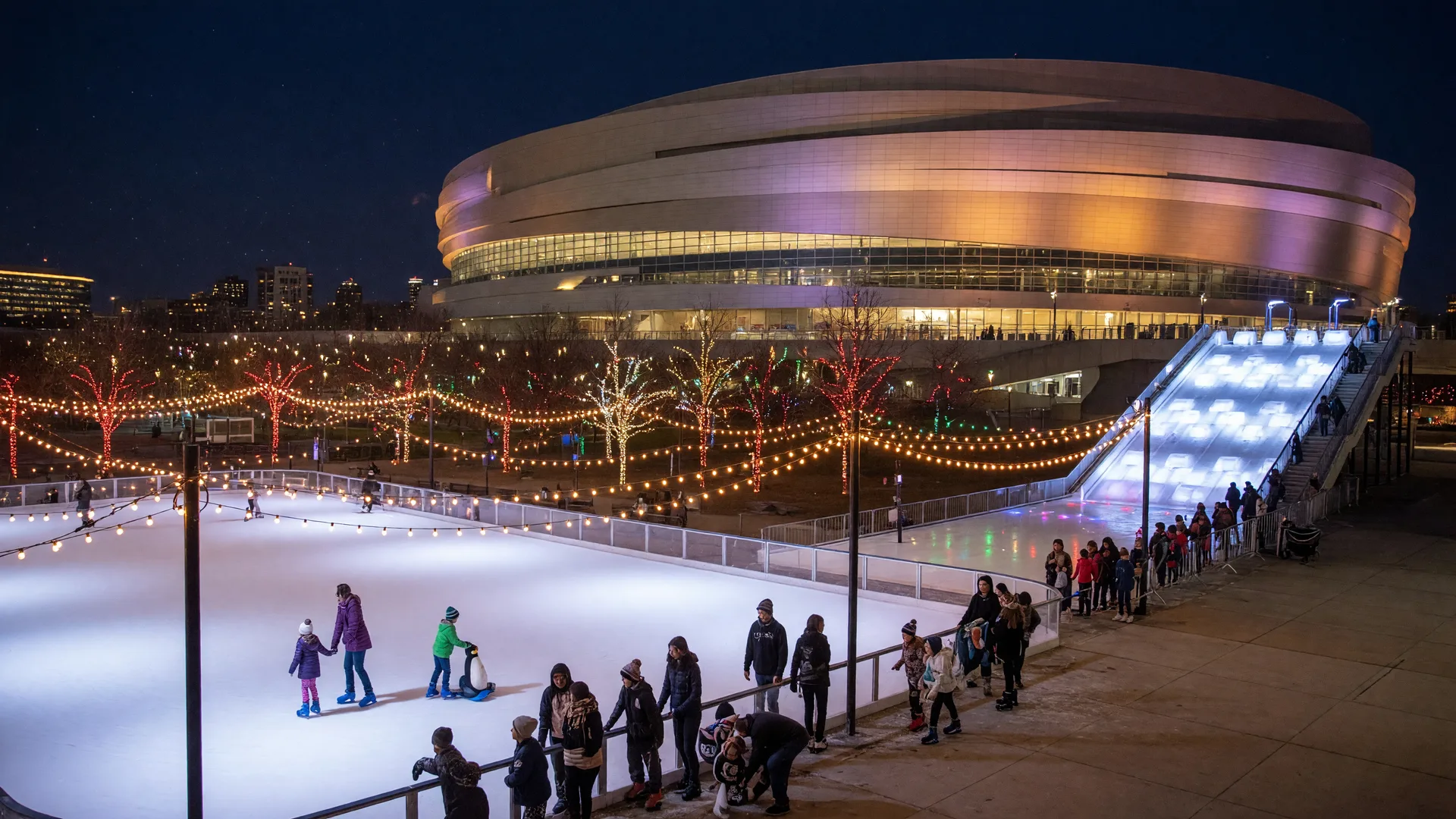 Outdoor ice skating rink on the BOK Center plaza in downtown Tulsa at night with glowing holiday lights and an ice slide