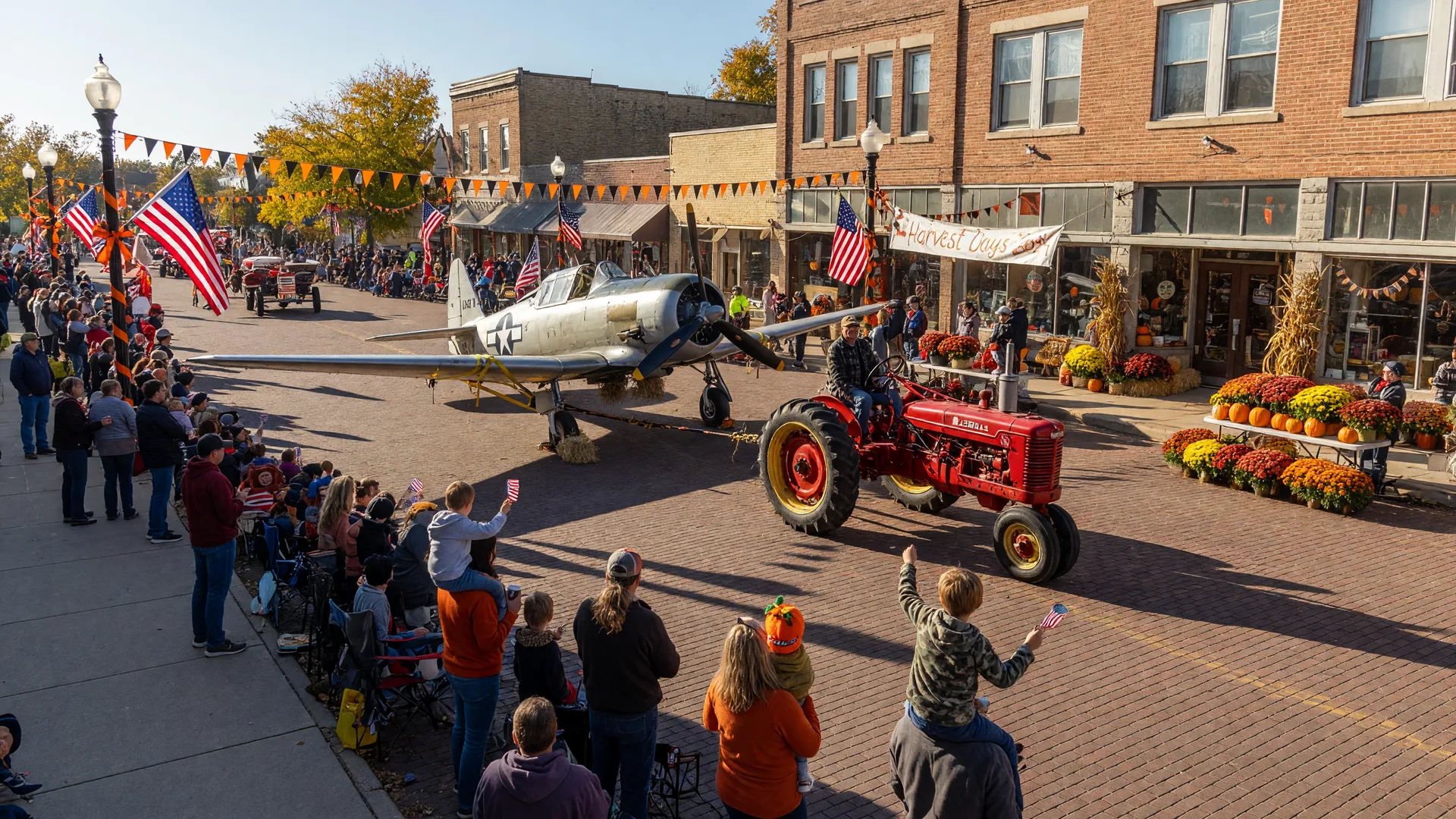 A vintage World War II airplane towed by an antique tractor down the brick main street of a small Ohio town during a fall festival parade, with waving crowds, flags, and autumn decorations