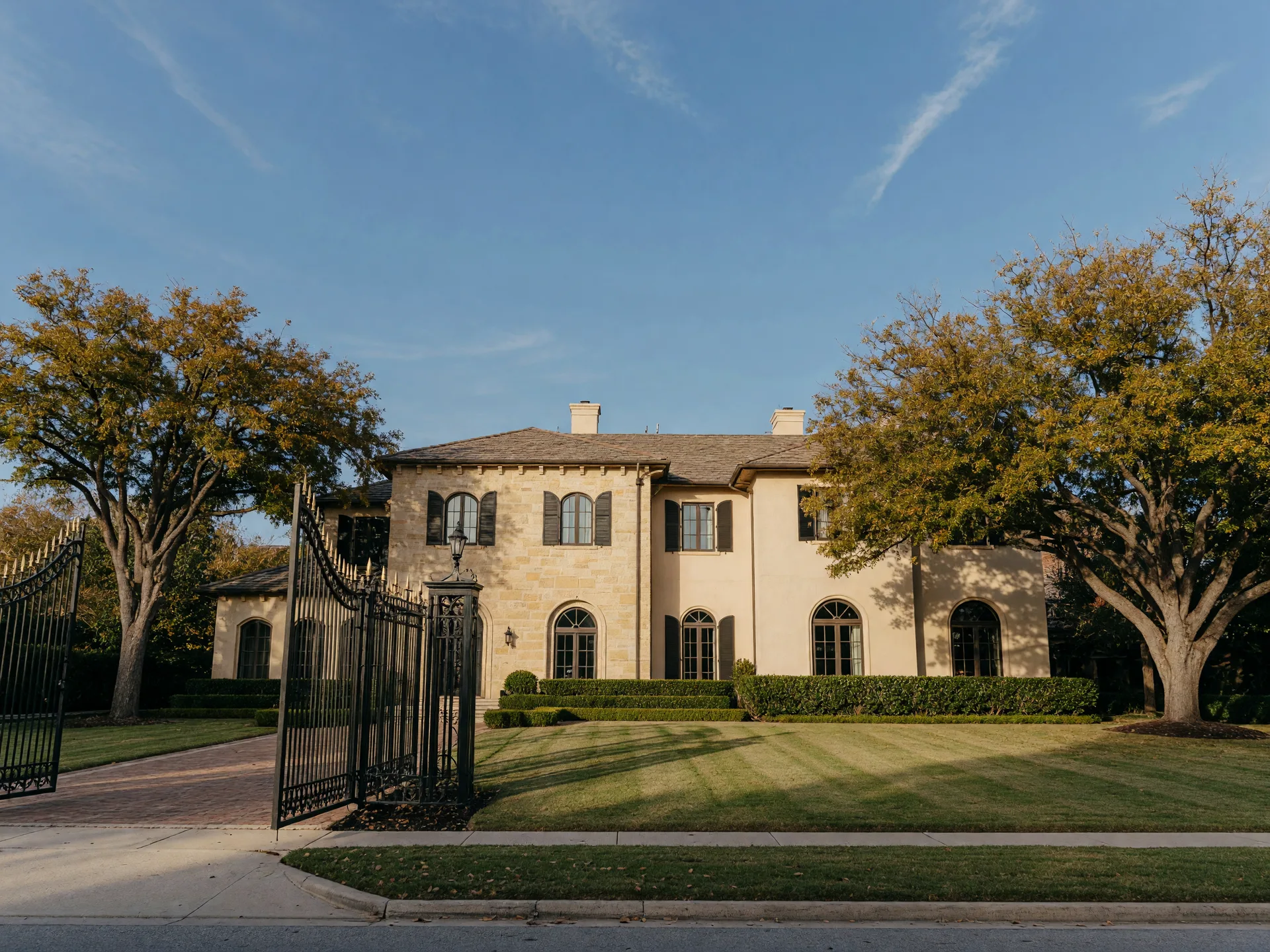 Luxury estate home in Dallas on a crisp mid-autumn afternoon, warm late-day light on stone and stucco with mature live oaks