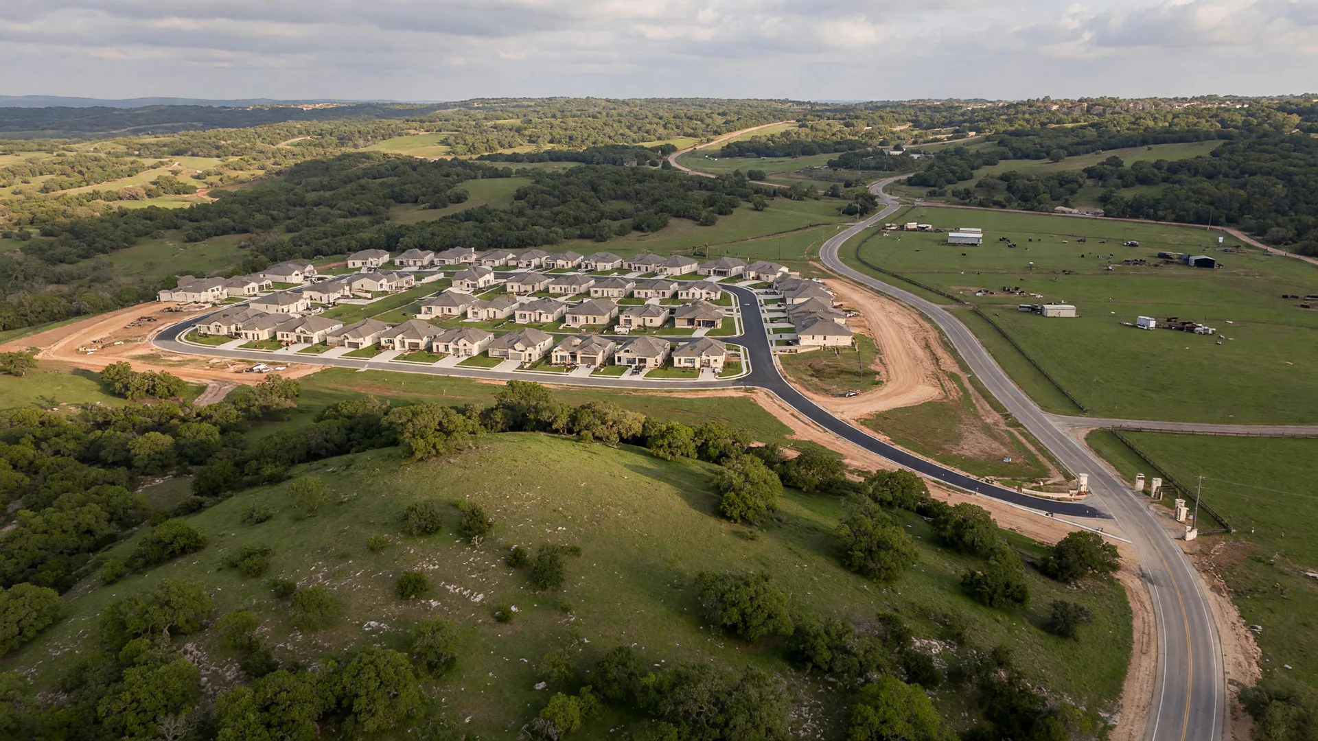 Aerial view of the Texas Hill Country showing new residential development alongside established ranchland, with rolling hills, live oaks, and winding country roads under golden afternoon light