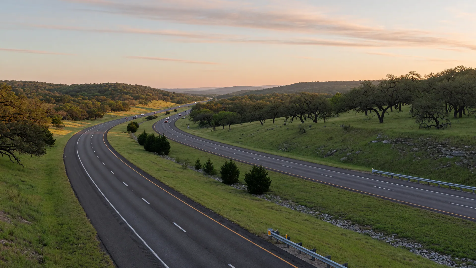 A Texas Hill Country divided highway curving through rolling green hills with live oaks at golden sunrise