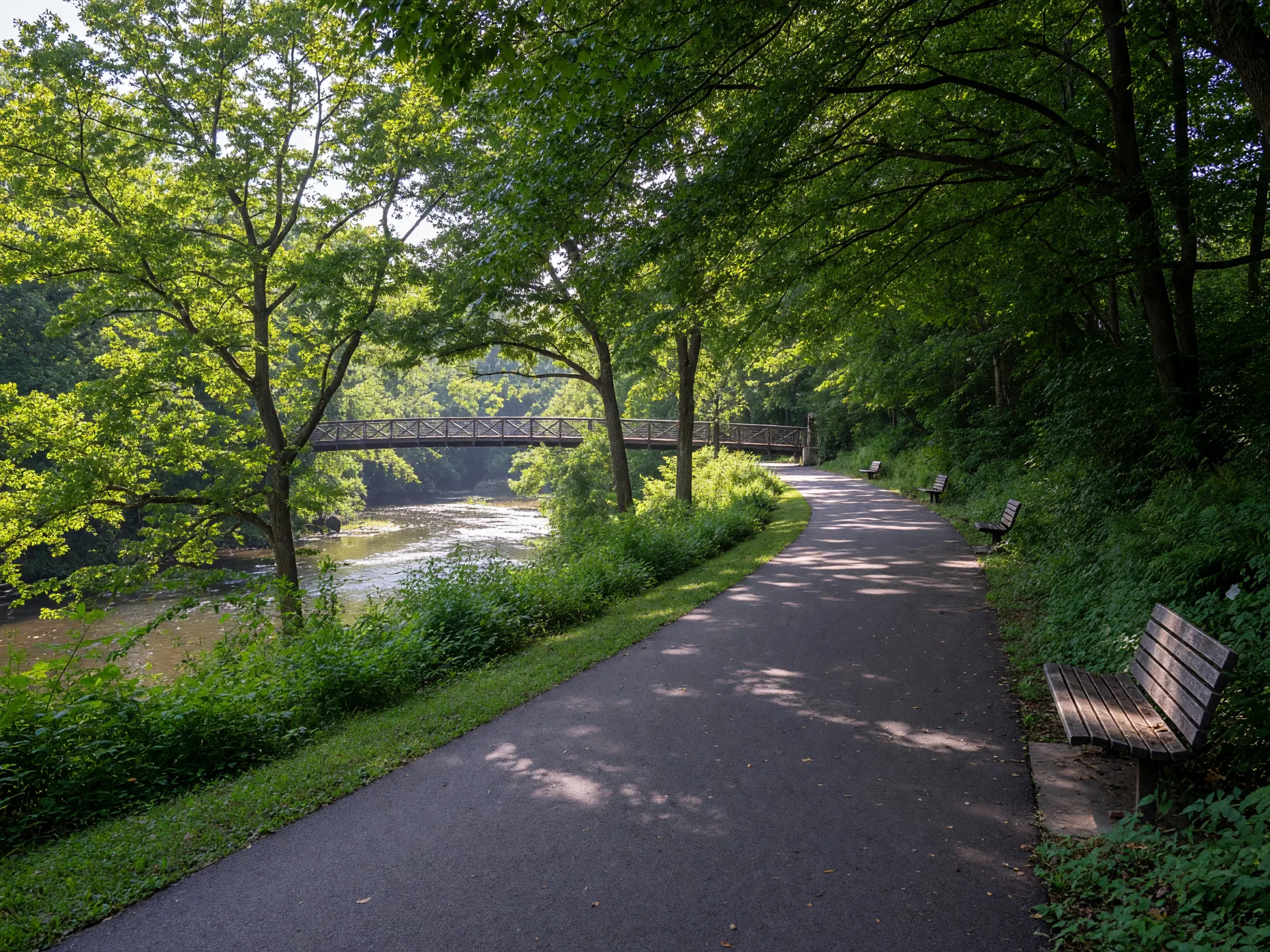 A wide paved greenway trail running beside a river through trees in Greenville, with a pedestrian bridge ahead
