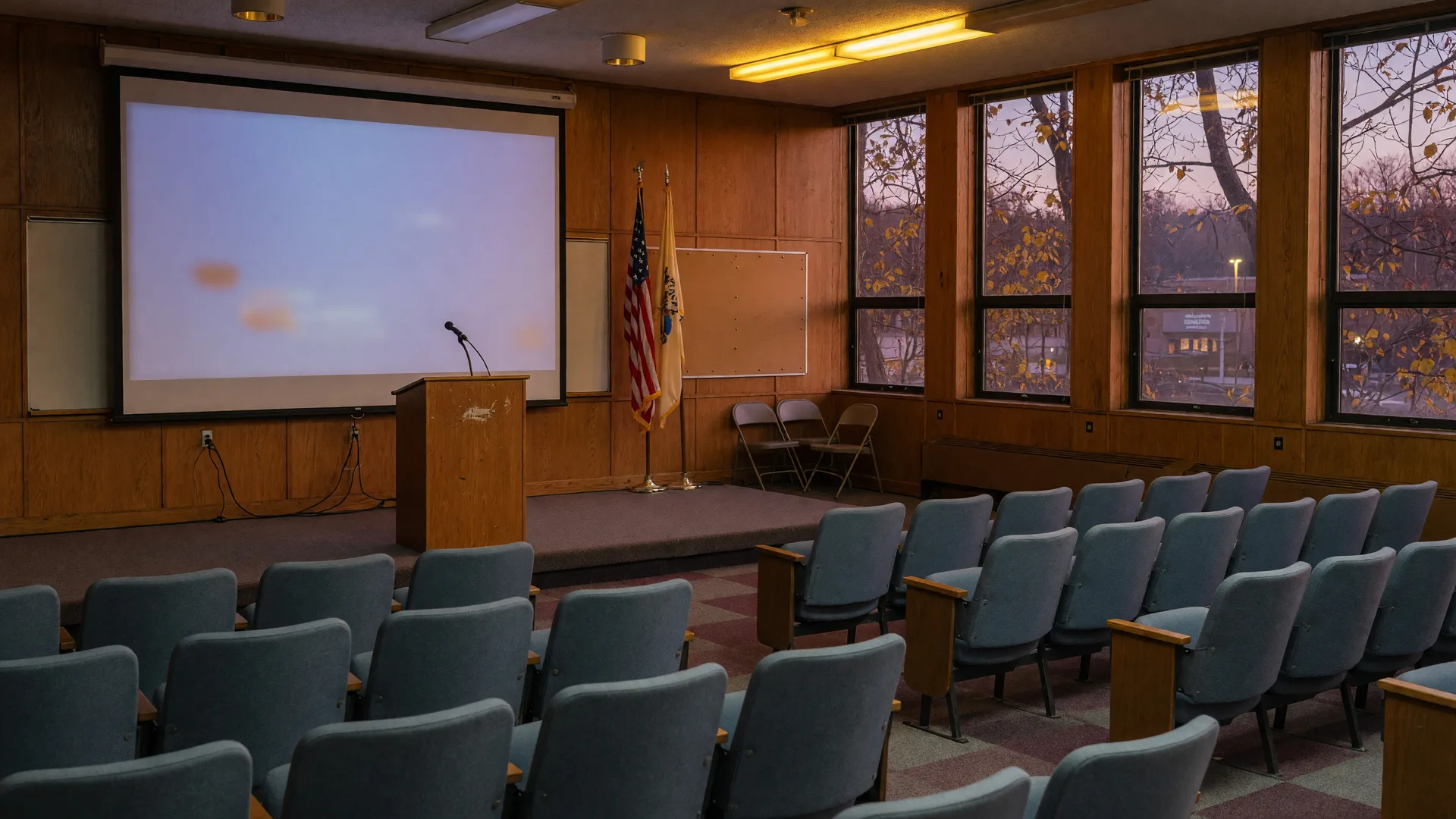 An empty school board meeting room in Montclair NJ on an autumn evening, rows of upholstered chairs facing a low stage with a microphone and a softly lit presentation screen, warm wood paneling