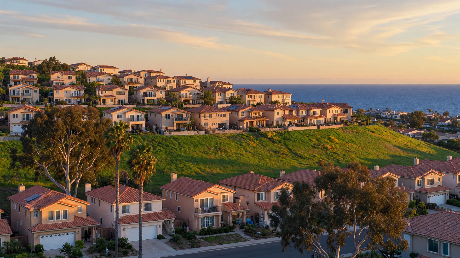 A North County San Diego hillside neighborhood at golden hour, tile-roofed family homes climbing a green hillside with a glimpse of the Pacific on the horizon