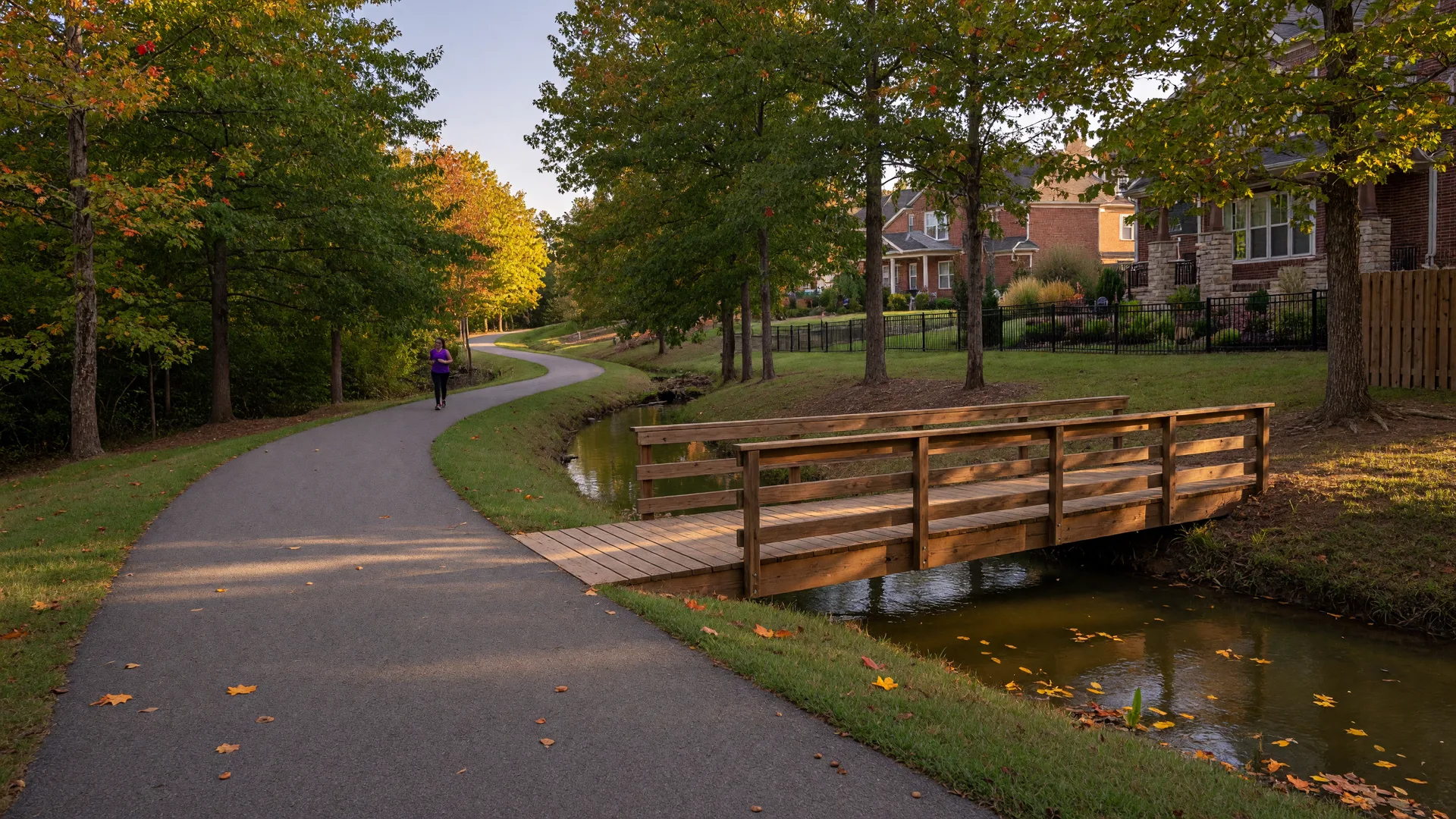 A paved greenway trail winding beside a creek in Huntersville, North Carolina, with a wooden pedestrian bridge and trees beginning to show fall color