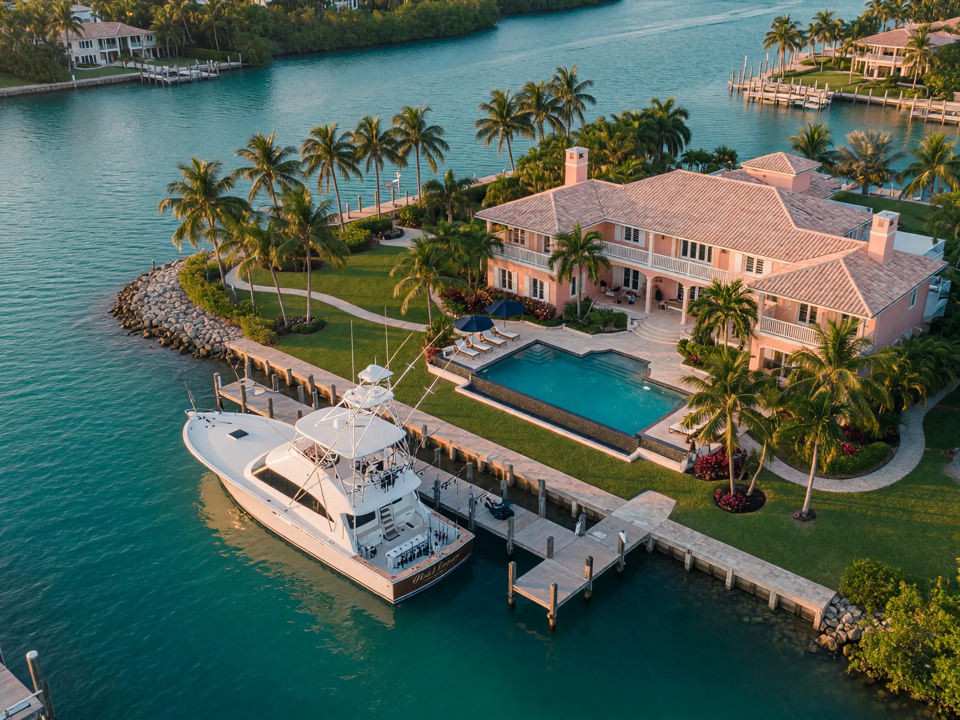 Aerial drone view of a luxury Florida Keys waterfront estate with a private dock, 50-foot sportfishing yacht, infinity pool, manicured tropical grounds, deep-water canal, golden hour lighting over turquoise water