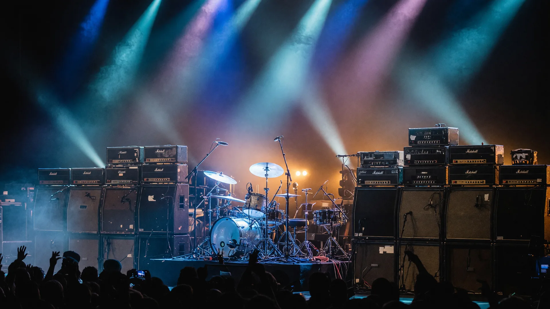 A rock concert stage with a wall of amplifiers and a drum kit lit by blue and white spotlights in thick haze