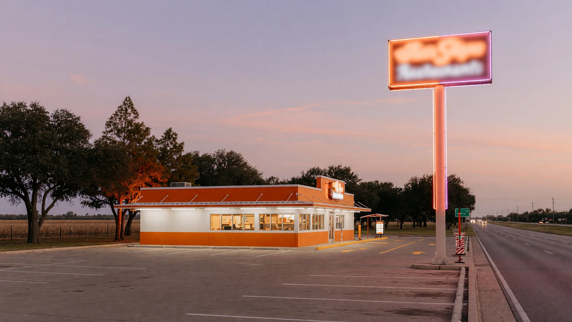 A modern quick-service restaurant with a drive-thru lane and illuminated sign pole on a wide commercial highway corridor in small-town Texas at dusk