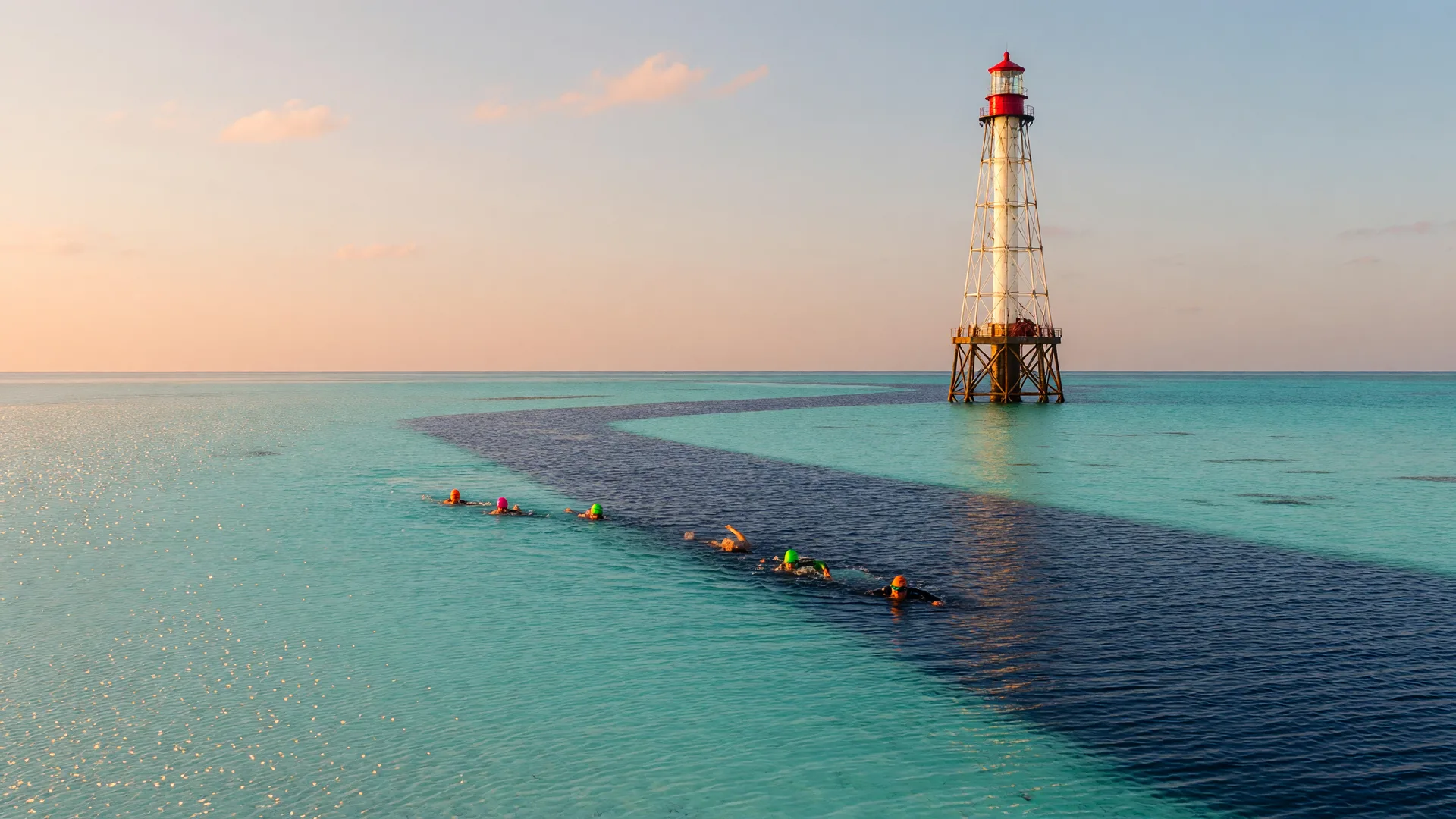 Alligator Reef Lighthouse rising from calm turquoise water at first light, with distant open-water swimmers in bright caps