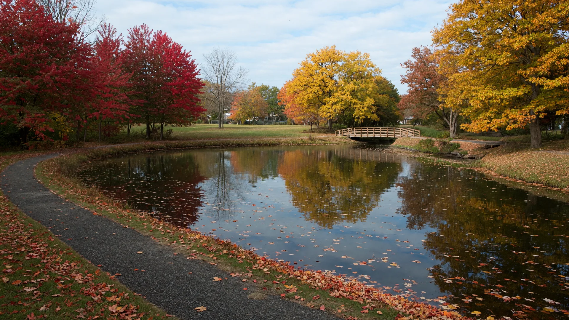 A small neighborhood pond in a suburban Essex County NJ park in late October, calm water ringed by red and gold autumn trees, with a walking path along the bank