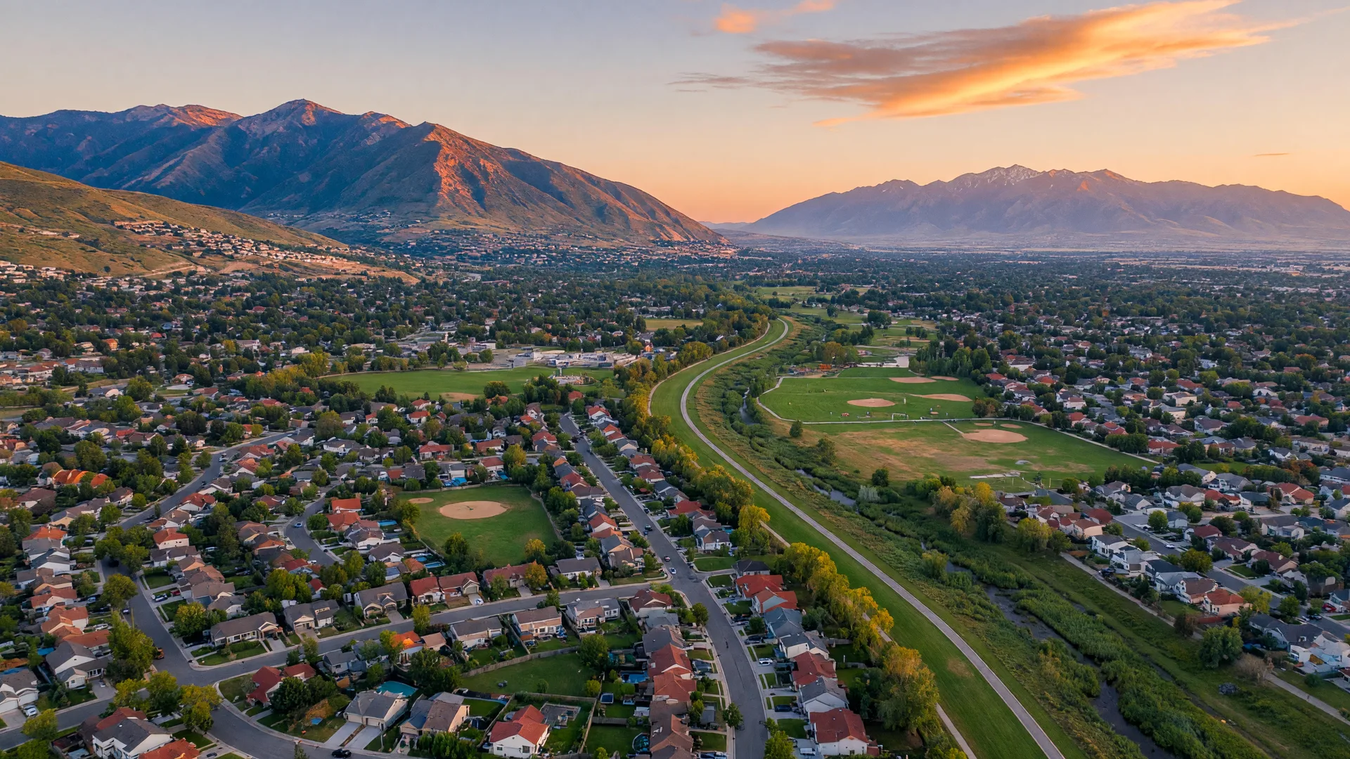 Aerial view of Riverton Utah at golden hour showing tree-lined neighborhoods, green parks, and the Jordan River Parkway Trail stretching toward the Oquirrh and Wasatch Mountains