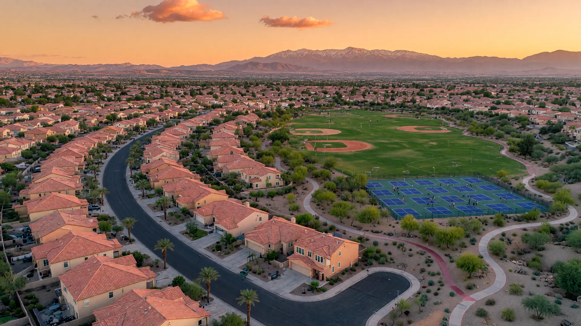 Golden-hour aerial of a Southwest Las Vegas master-planned community with red-tile roofs, a regional park, sports fields, and the Spring Mountains