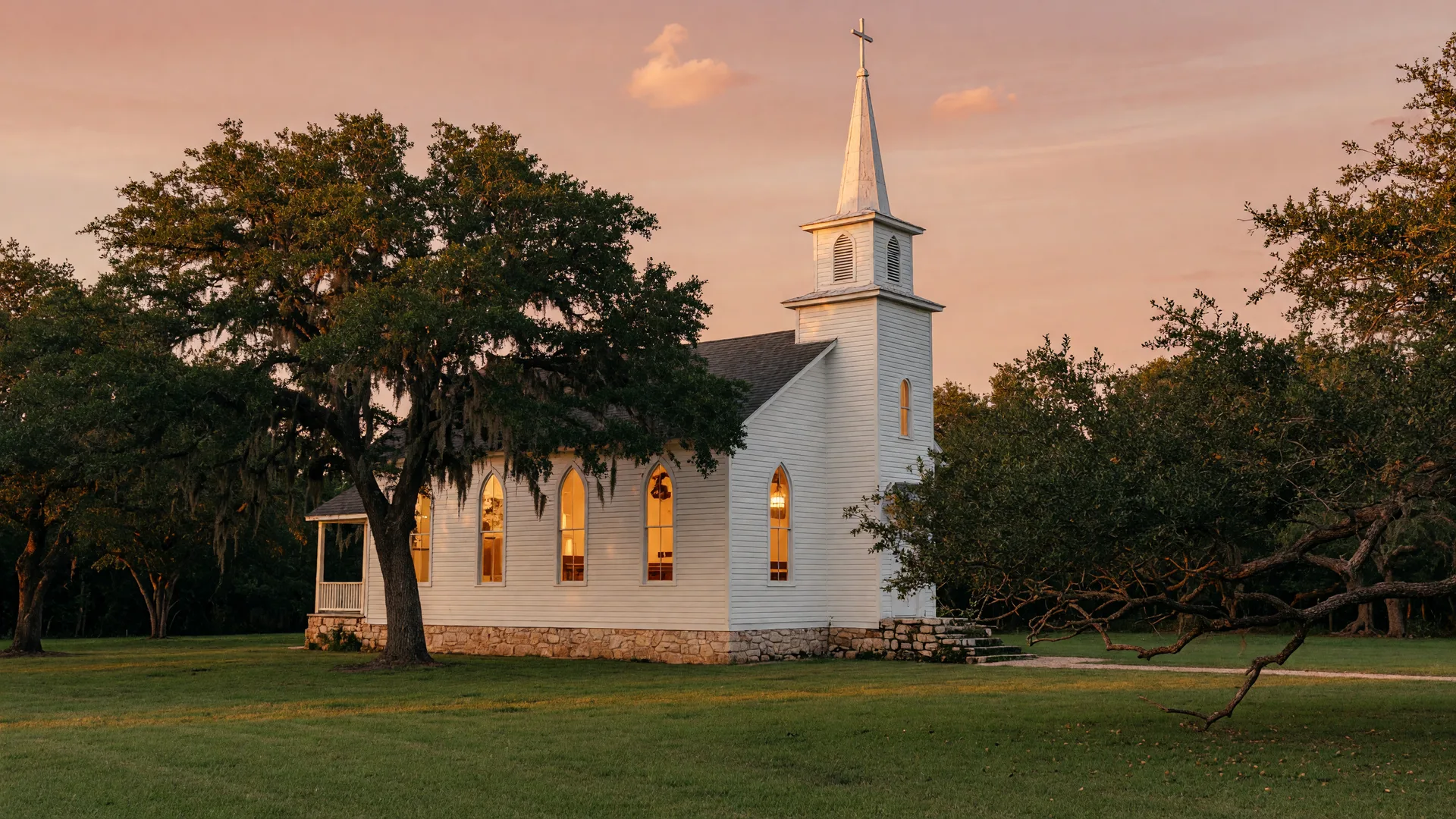 A white frame Texas Hill Country chapel with a tall steeple and glowing arched windows at golden hour, framed by live oaks