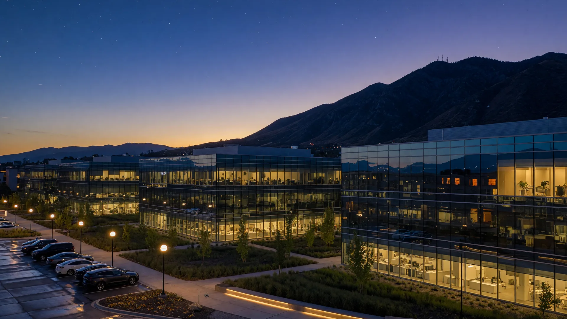 A modern tech office campus along Utah's Wasatch Front at dusk, glass buildings glowing with warm interior light against the mountain silhouette.