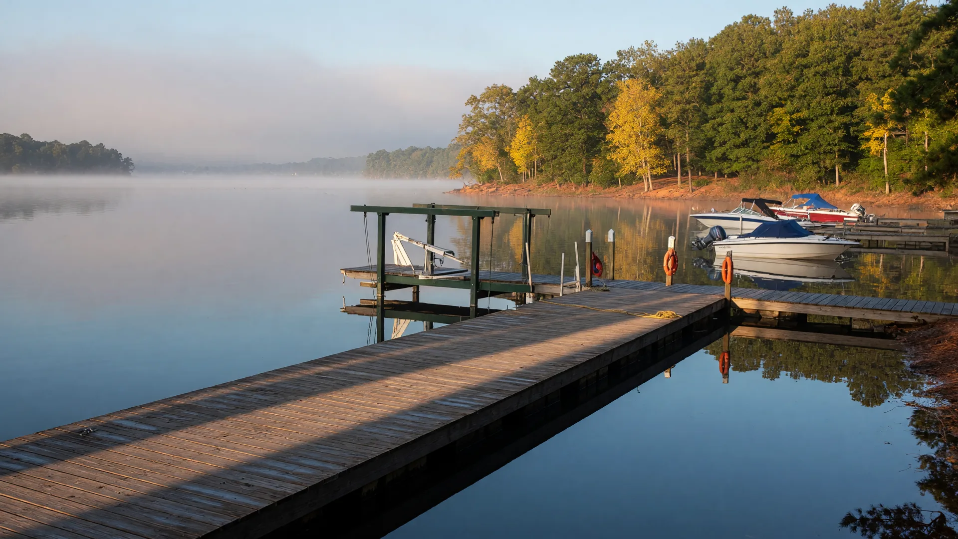 A long wooden dock and boat lift reaching into calm September water at Lake Norman, with the first hints of autumn color in the shoreline trees and soft morning light