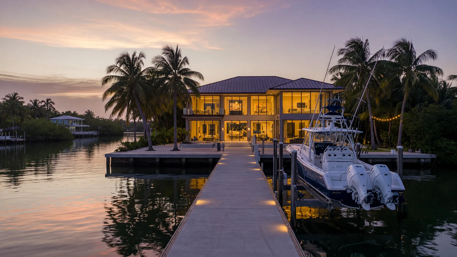 A luxury Florida Keys waterfront estate at dusk viewed from the canal, an elevated modern home with impact glass glowing warm amber, a private concrete dock with a boat lift and a sportfishing yacht, a deep-water canal reflecting a champagne and lavender sky