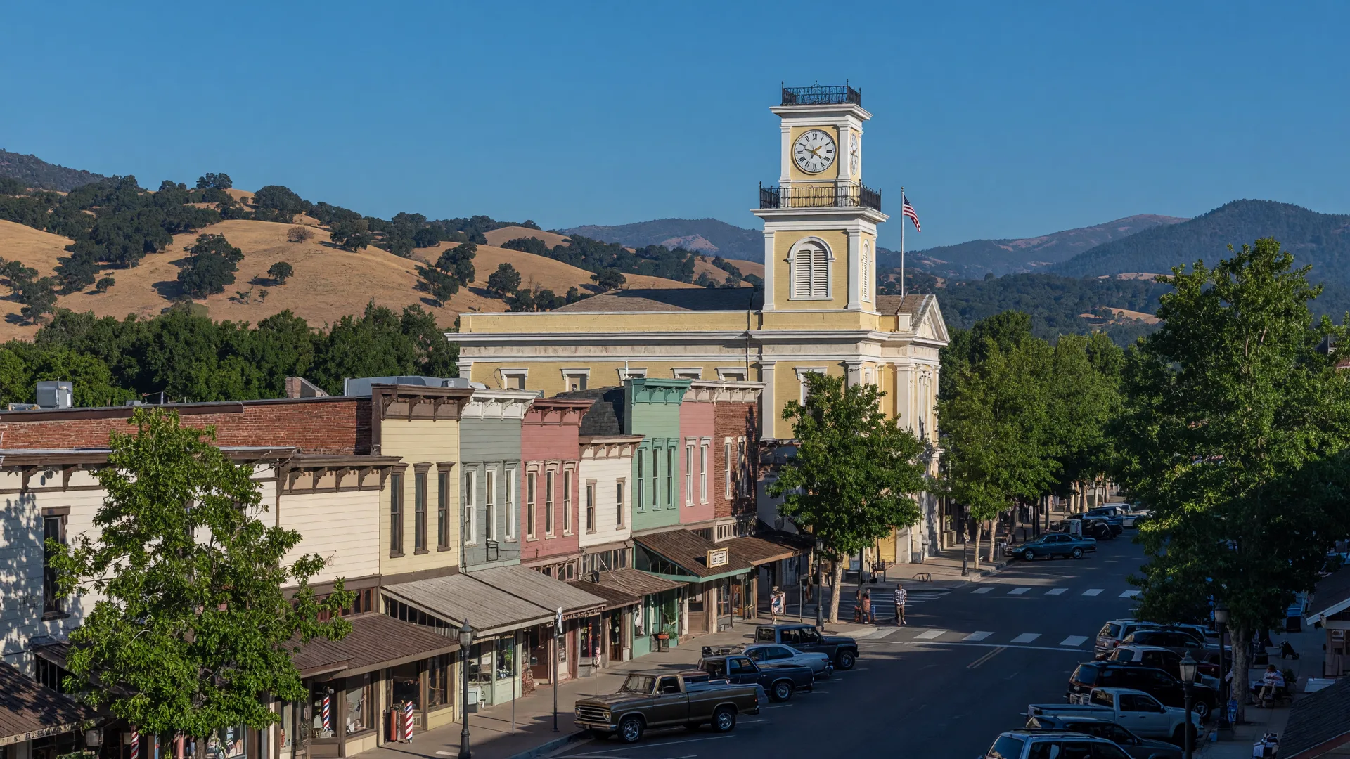 Historic Mariposa, California main street on a bright summer morning with Gold Rush-era storefronts, the Mariposa County Courthouse clock tower against a blue sky, and Sierra Nevada foothills in the background