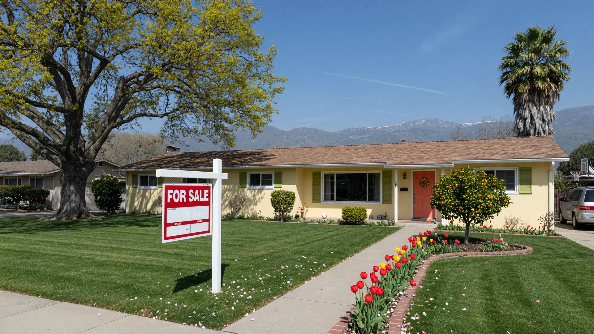 A well-presented single-story ranch home in a Merced County neighborhood on a sunny spring afternoon with a for-sale sign in the front lawn, mature oak and palm trees, and the Sierra Nevada foothills on the horizon