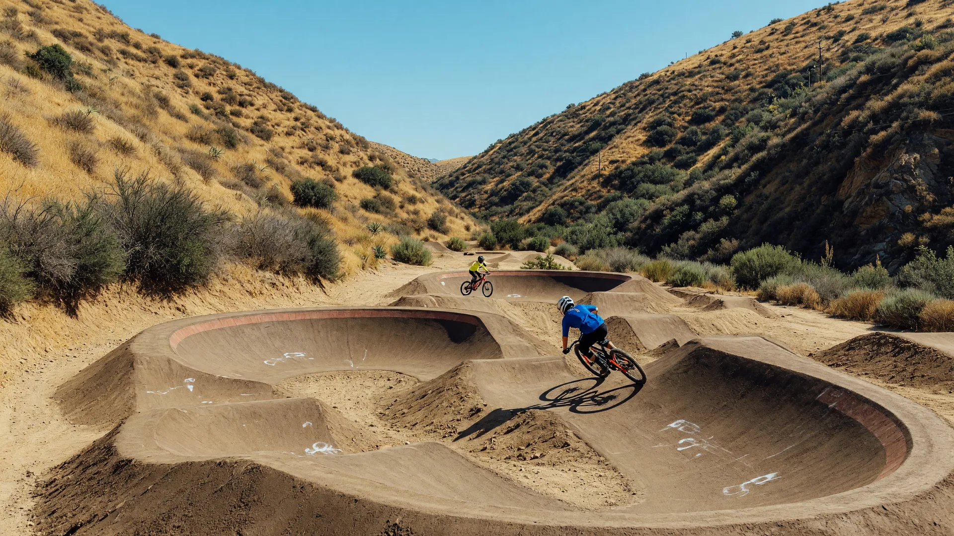 A modern mountain bike skills park in a Southern California canyon with a sculpted dirt pump track and jump lines