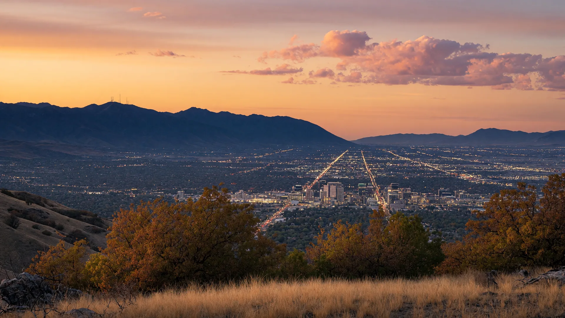 The Salt Lake Valley seen from the east bench foothills at sunset, neighborhood lights spreading across the valley floor with the Oquirrh Mountains on the western horizon.