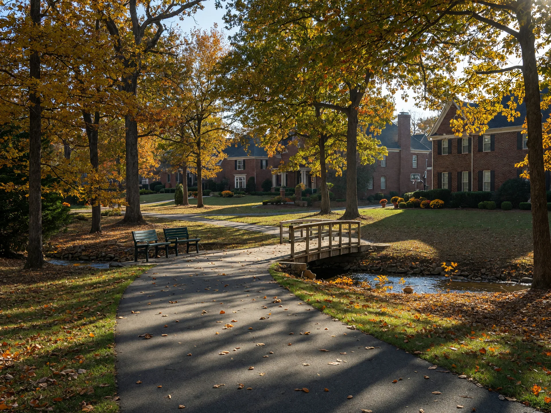 Tree-lined greenway trail in Farragut, Tennessee with a wooden footbridge, park benches, and upscale brick homes visible through the autumn trees