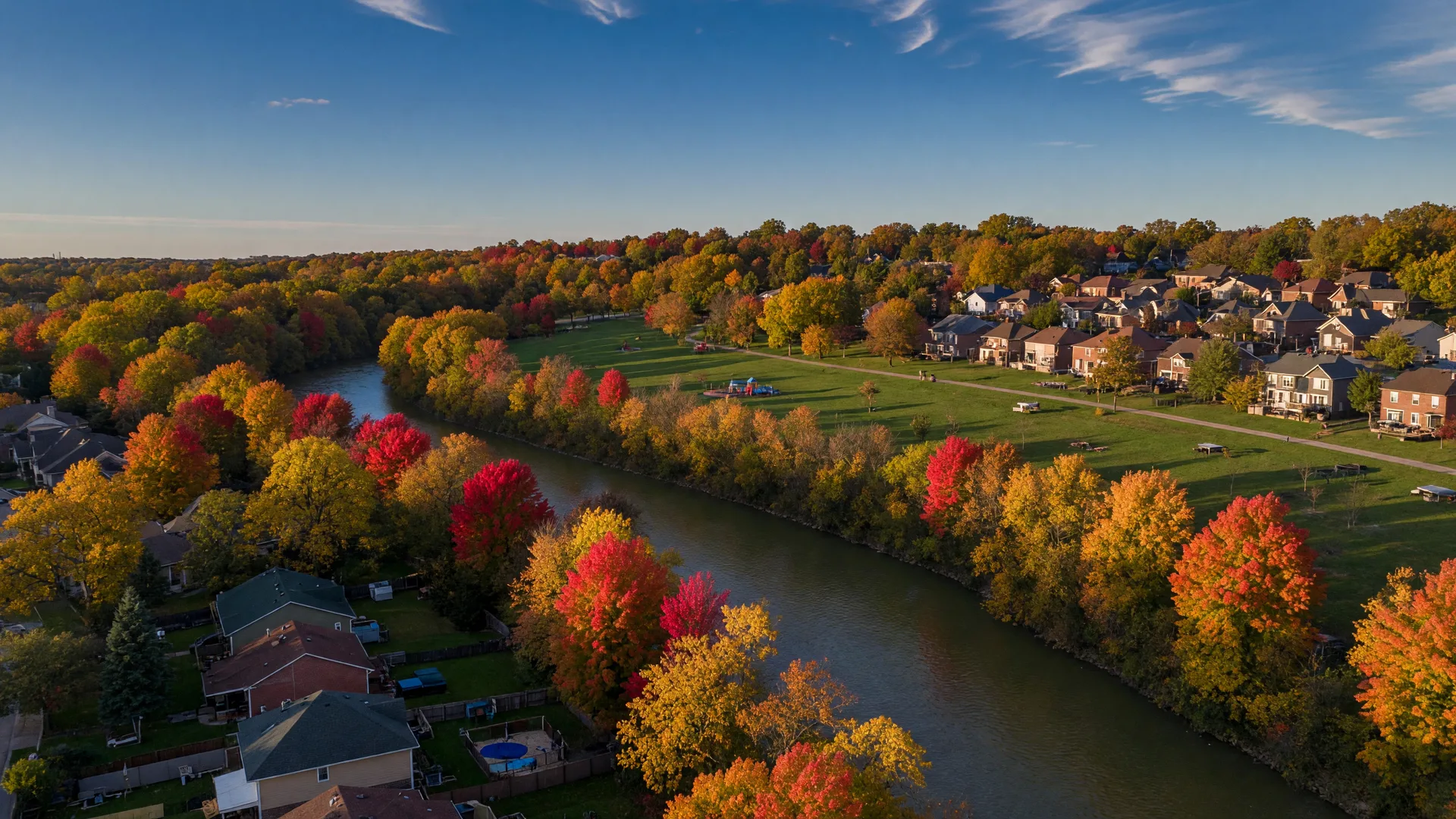 An early-autumn aerial view of Beavercreek, Ohio with the Little Miami River curving through a canopy of orange and gold trees, suburban rooftops and green parkland in warm late-afternoon light