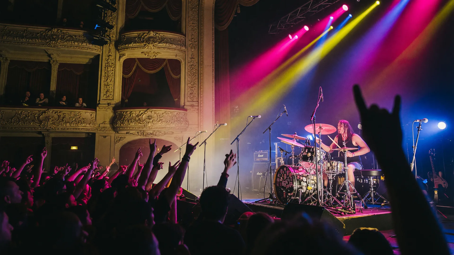 A hard rock concert at a grand theater with dramatic magenta, yellow, and blue stage lights cutting through haze, a drummer behind a large kit, anda silhouetted crowd with raised hands
