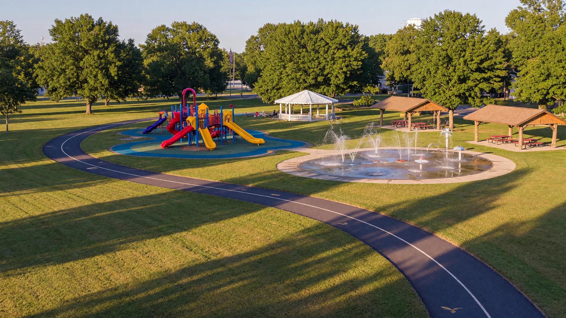 Well-kept town park with a playground, splash pad, and paved trail through mature trees in late-summer light