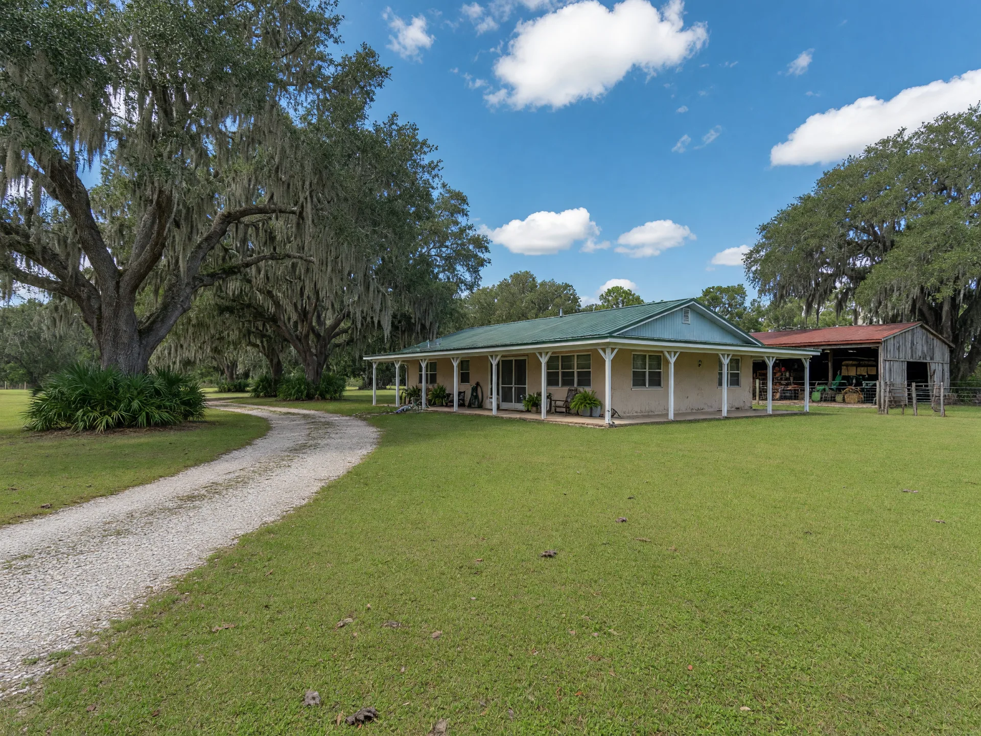A single-story ranch-style acreage home with a metal roof and covered porch, a pole barn, and a gravel drive lined with live oaks under a blue Florida sky