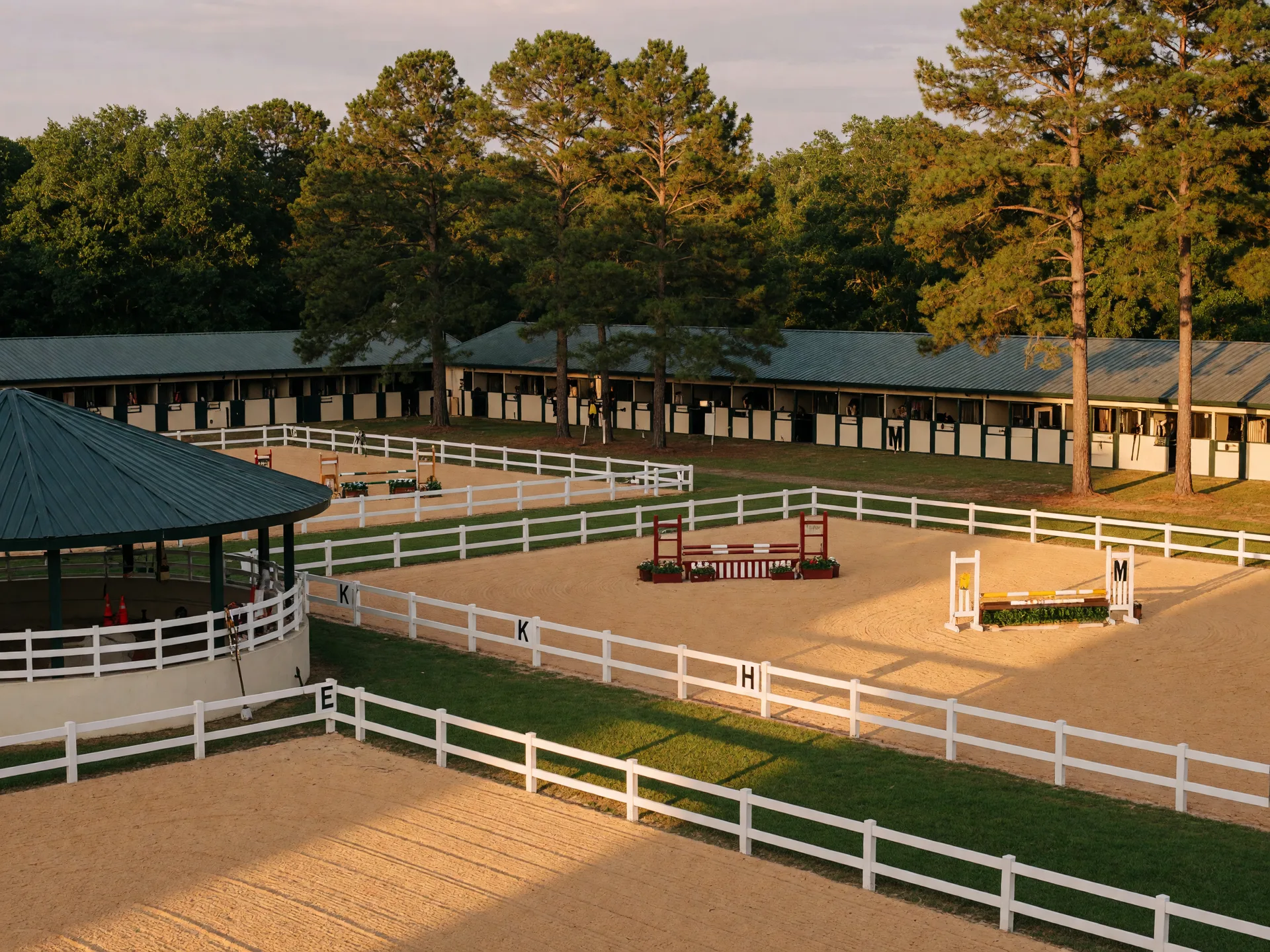 White-fenced equestrian arenas and training facilities at an Aiken horse venue in late afternoon golden light with mature pine trees and stable complex