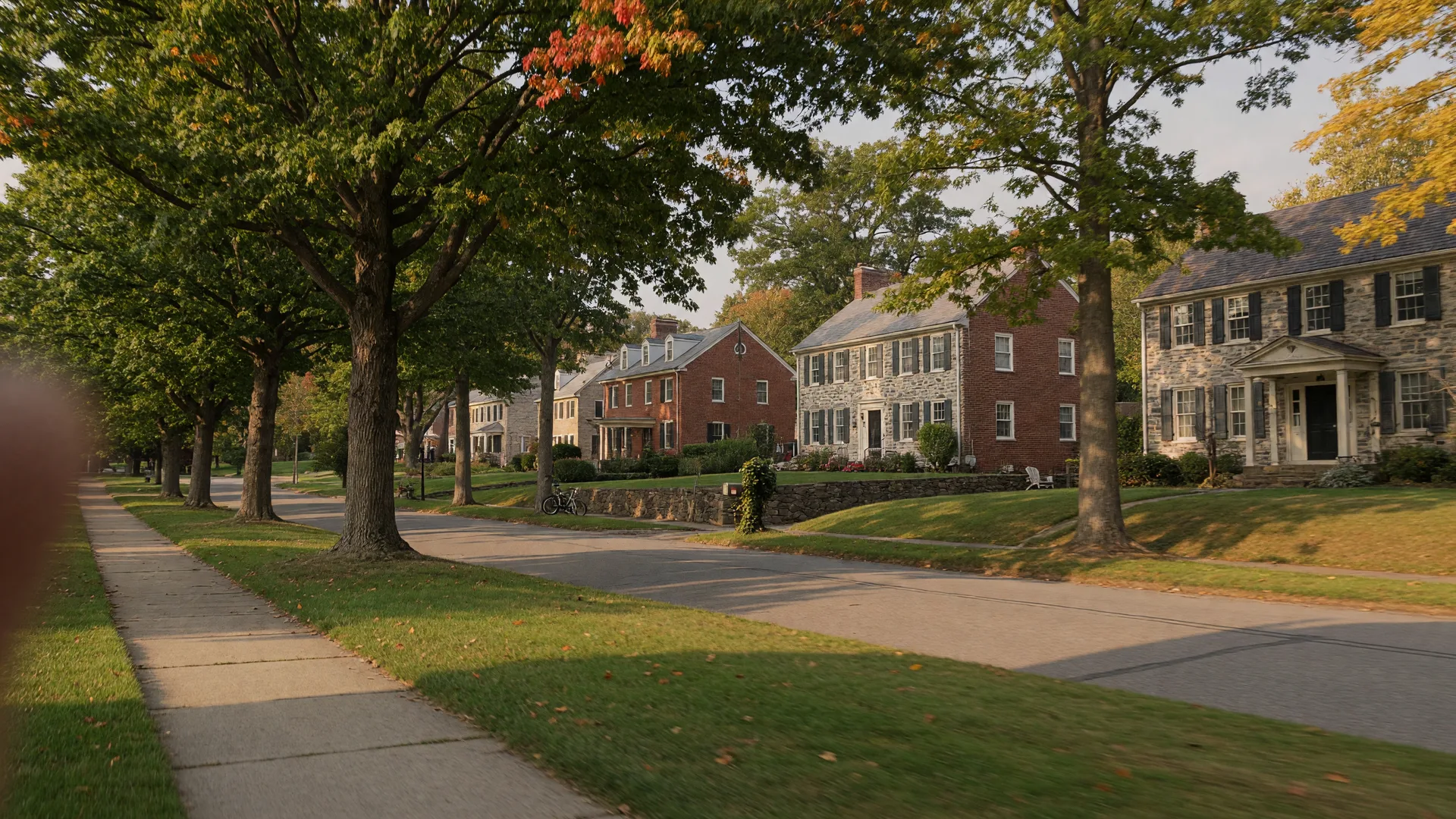 A tree-lined residential street in Rydal PA in mid-September with golden late-afternoon light across stone and brick homes under mature oaks with the first hints of autumn color