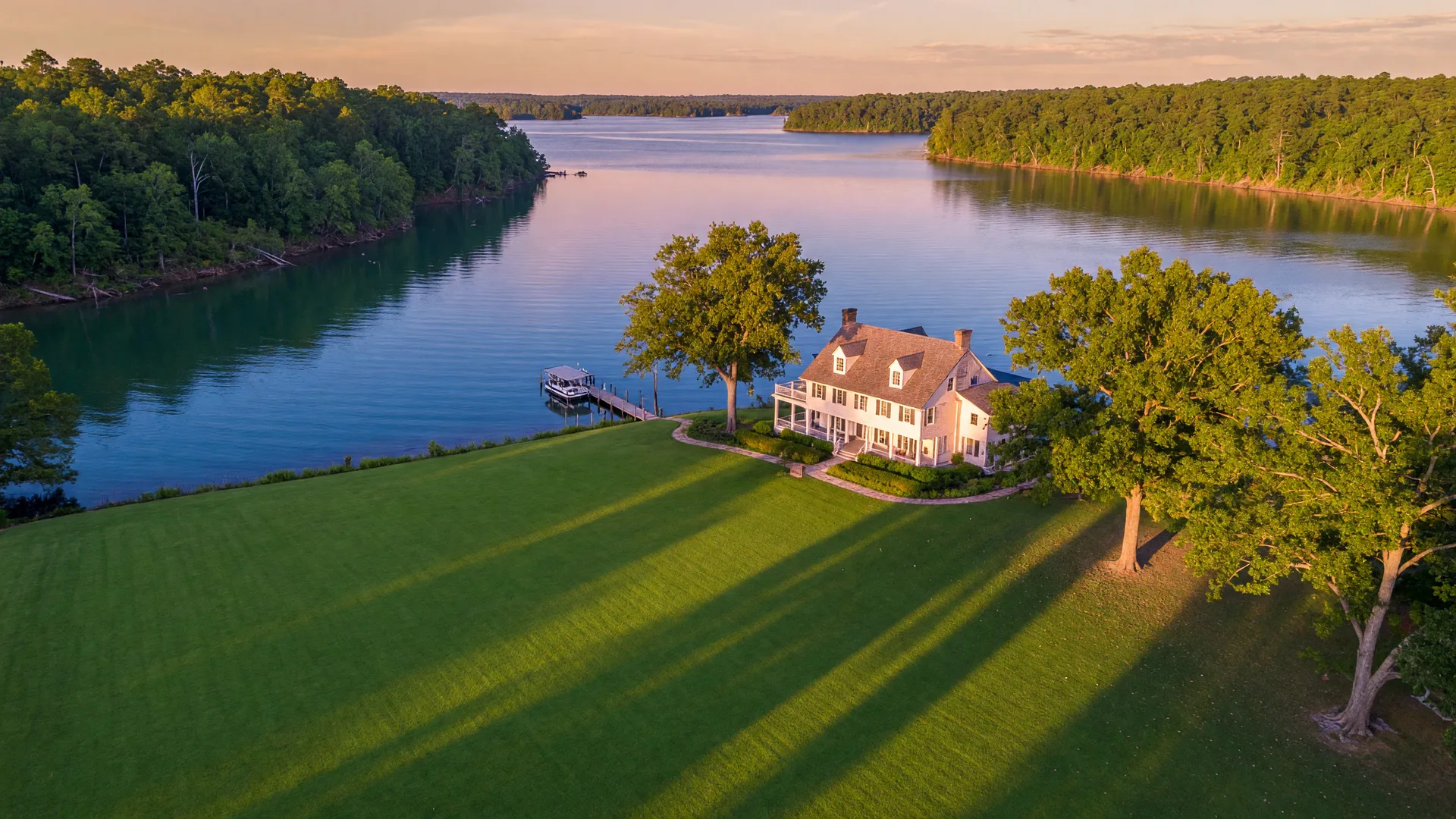 Aerial drone view of the waterfront estate at 257 Methodist Park Lane at golden hour, with flat green lawn, mature pecan trees casting long shadows, white farmhouse, and blue lake beyond