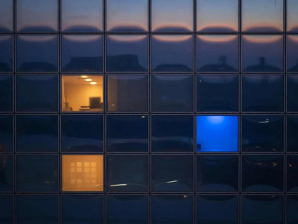 Glass skyscraper facade at dusk with one electric blue window among warm lights