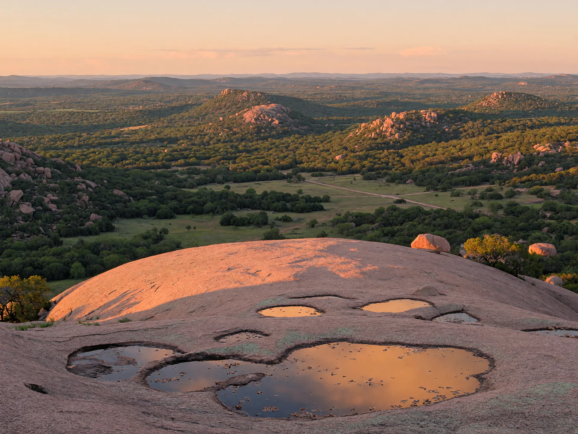 Wide panoramic view from the summit of Enchanted Rock showing the pink granite foreground with shallow vernal pools and the Texas Hill Country stretching to the horizon in golden light