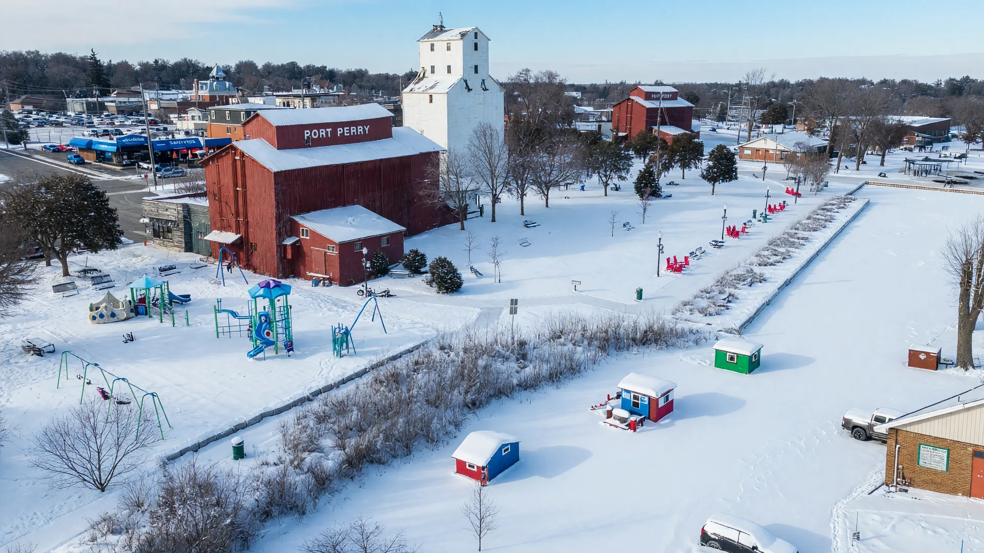 The Port Perry waterfront in winter: the historic red-brick mill and white grain elevator rising beside a snow-covered frozen Lake Scugog, with ice-fishing huts on the white ice and snow-dusted trees along the shore