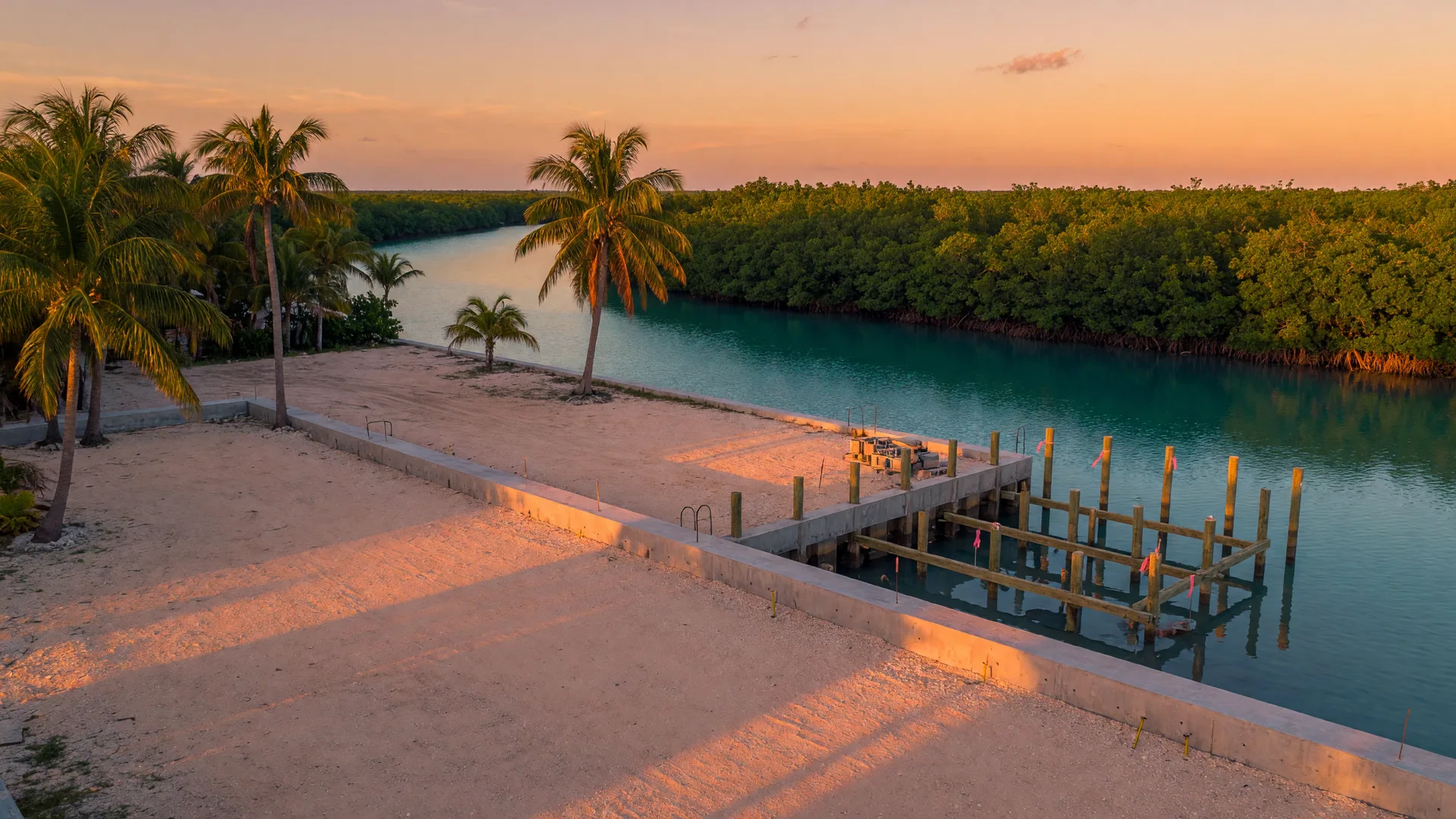 A rare buildable waterfront homesite in the Florida Keys at golden hour, an elevated cleared lot with a concrete seawall along a deep-water canal, preserved mangroves, and the start of pilings for a raised foundation