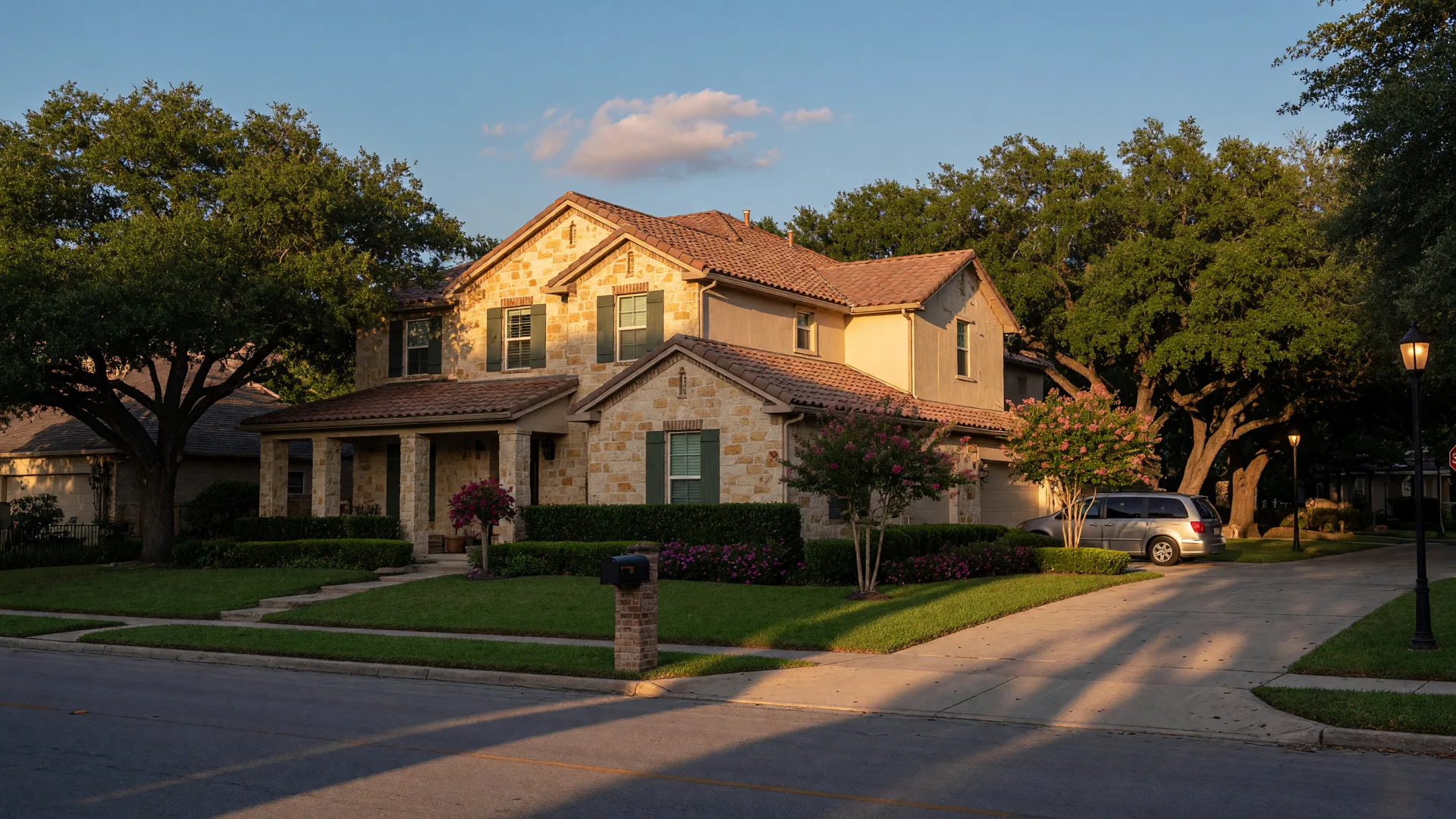 Two-story stone-and-stucco family home in Stone Oak, a master-planned San Antonio real estate community, at golden hour with live oaks and a greenbelt behind it