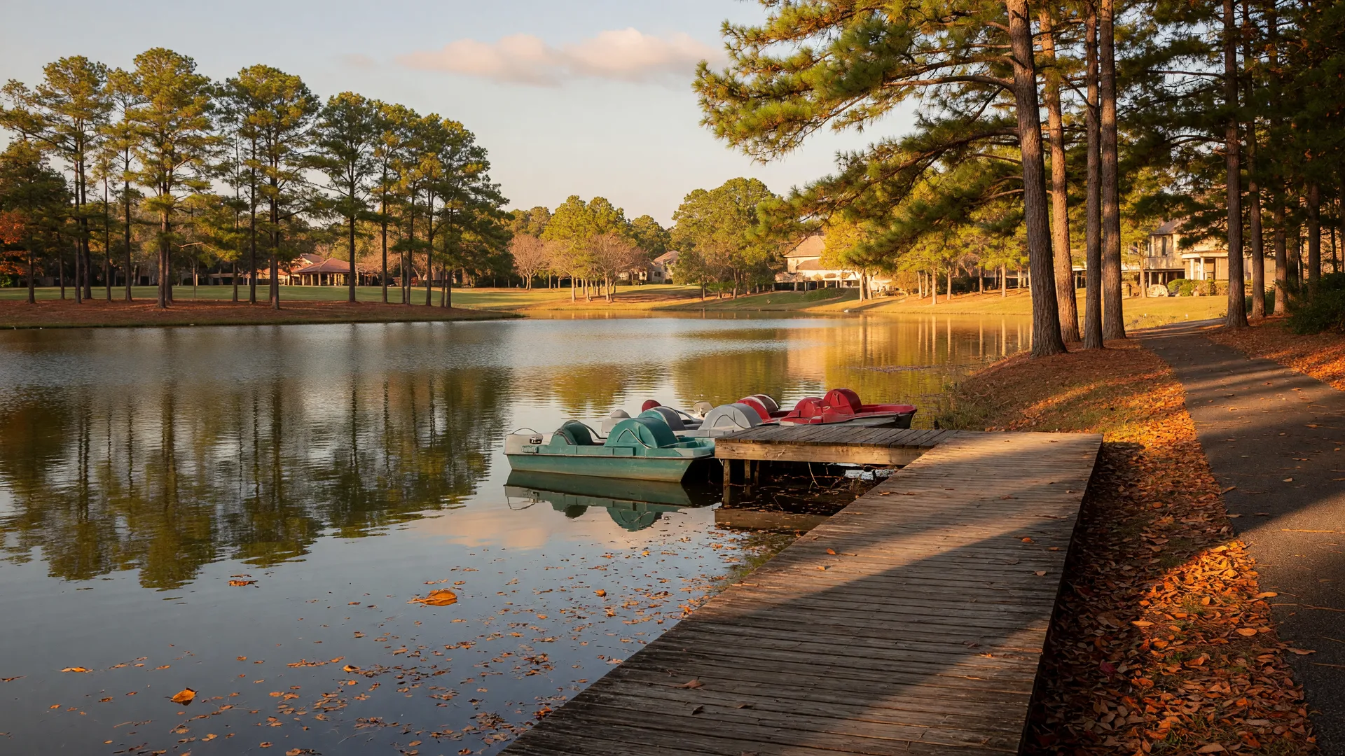 A five-acre community lake in a Texas master-planned neighborhood in mid-October, golden light through loblolly pines, a wooden boardwalk, and paddle boats at the dock