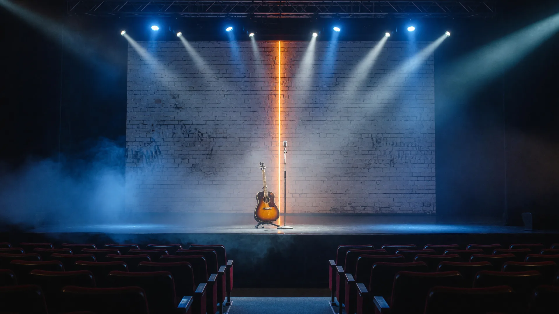 A massive white brick stage wall with a seam of warm light, blue and white beams cutting through haze over a microphone and acoustic guitar on a dark stage