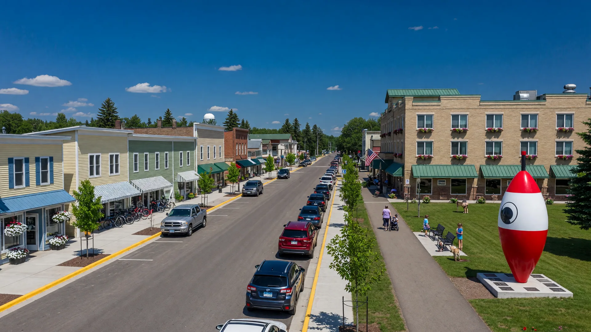 A tidy small-town main street with new shops and a fishing bobber monument beside a trail