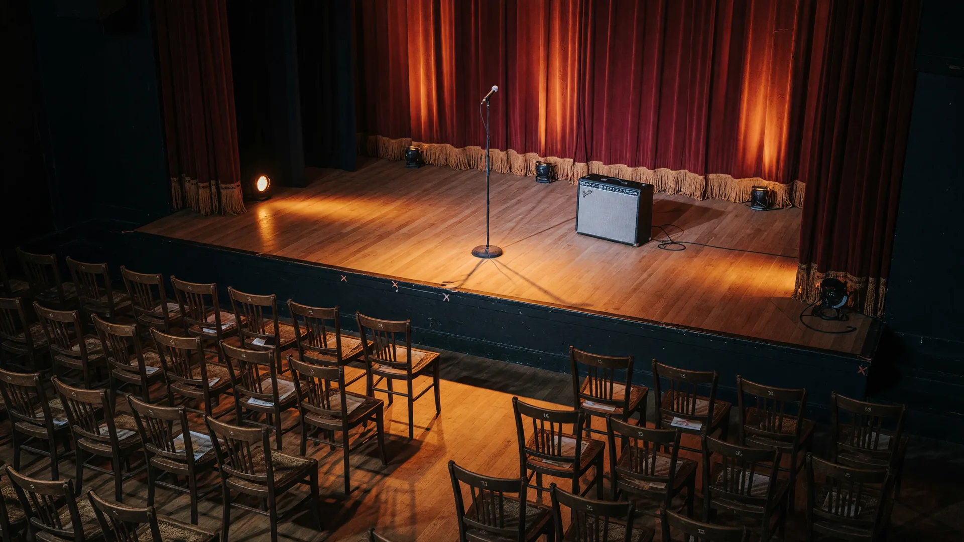 An intimate indoor music venue stage with warm amber stage lights, microphone stand, guitar amp, and red curtain backdrop