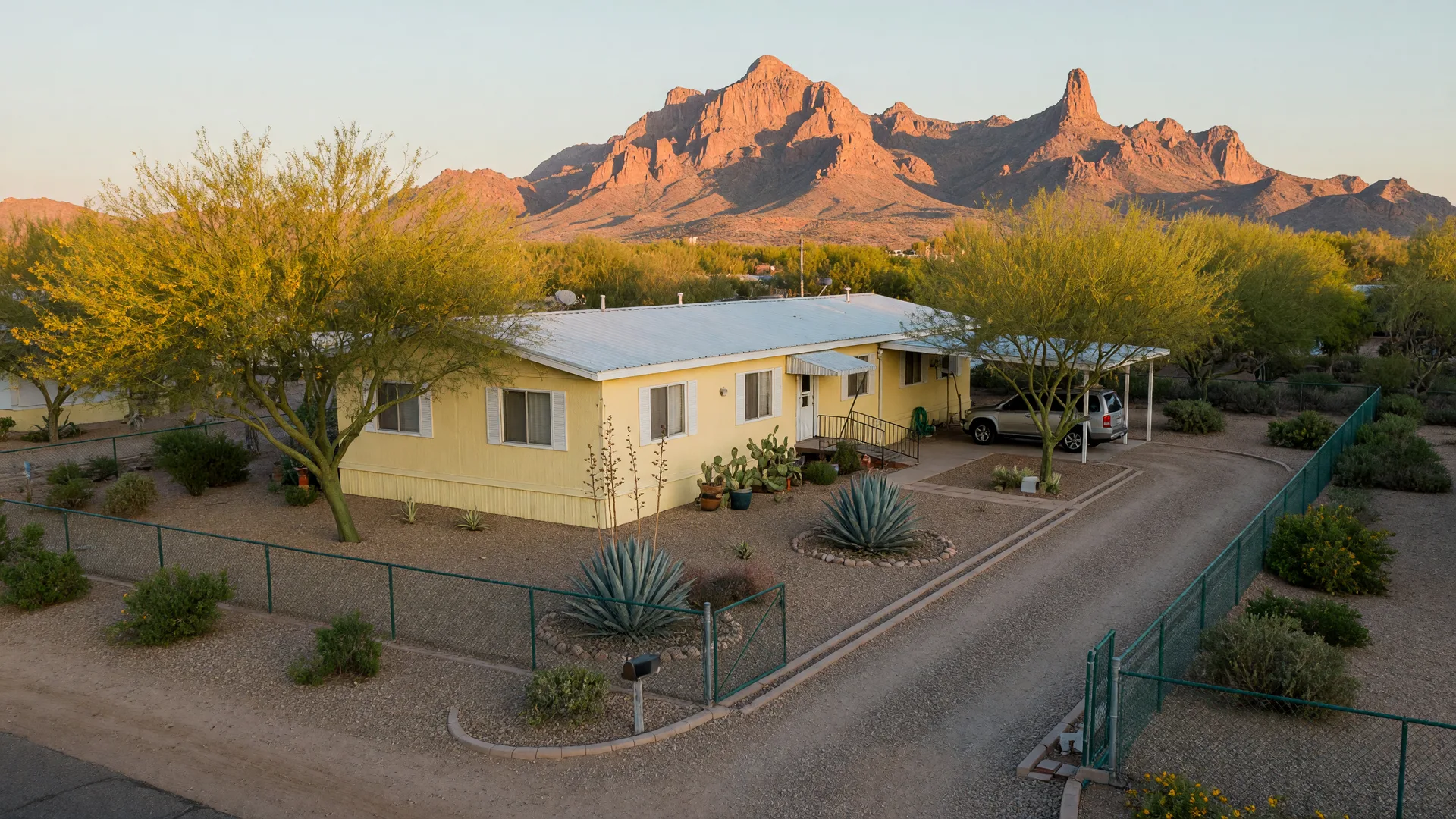 A single-story land-owned manufactured home with a metal roof and covered carport on its own fenced desert lot in Mesa, Arizona at golden hour, with palo verde trees and the Superstition Mountains on the horizon