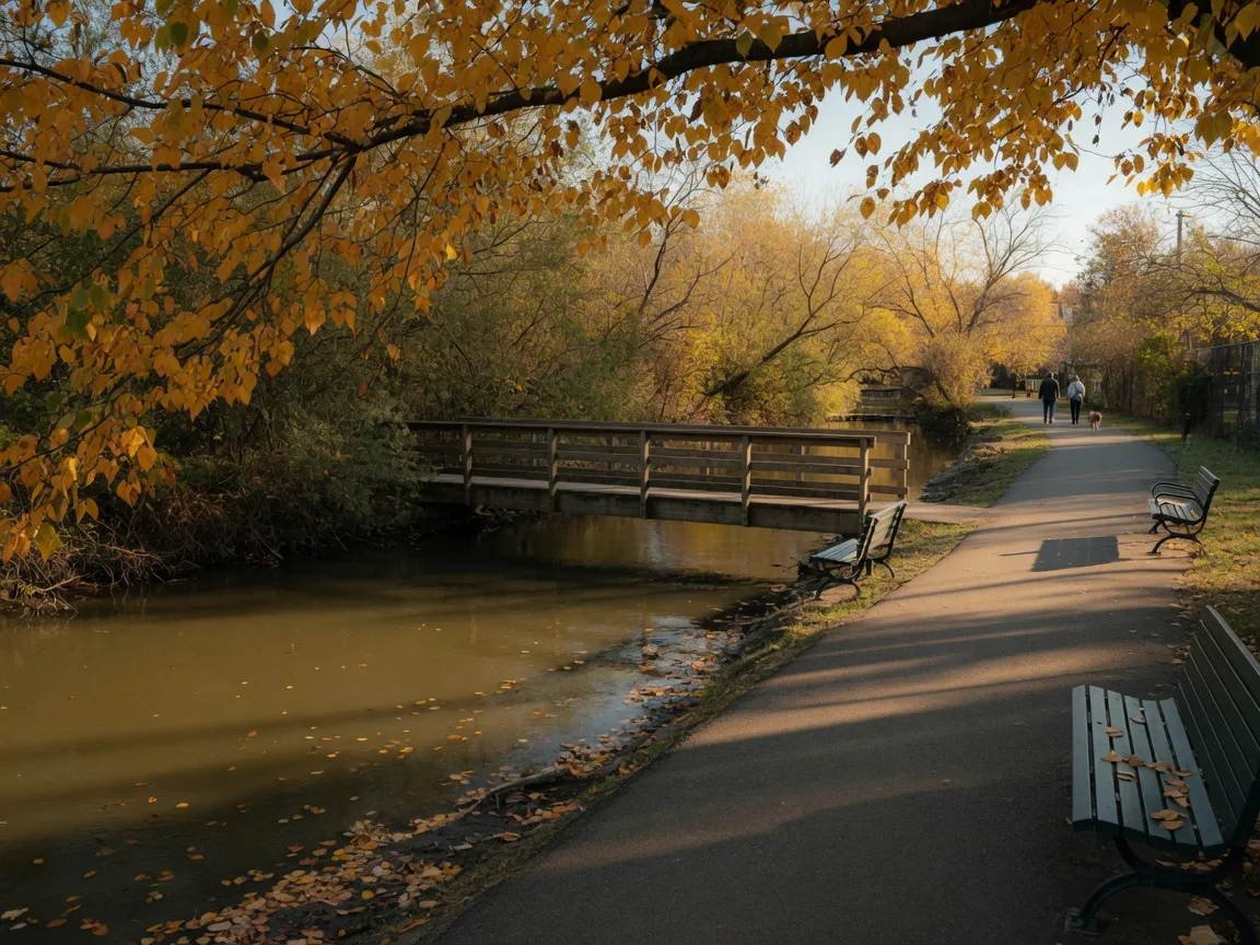A calm New Jersey riverside park in late October, a paved walking path along a slow creek under golden autumn foliage, with a wooden footbridge and benches in warm light