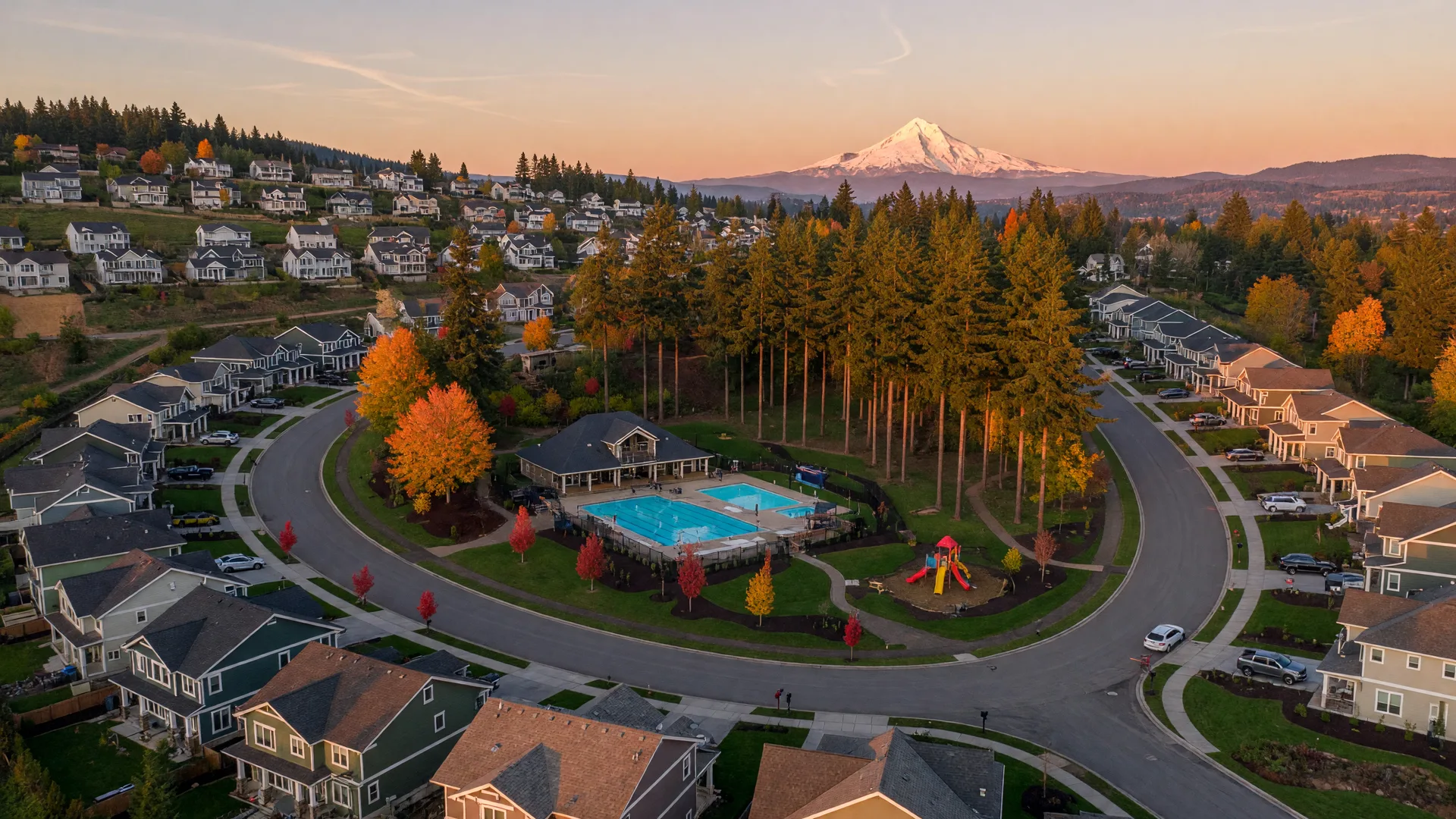 Elevated golden-hour view over a master-planned Happy Valley hillside community with curving streets of craftsman homes, a park with a pool, and Mount Hood on the horizon
