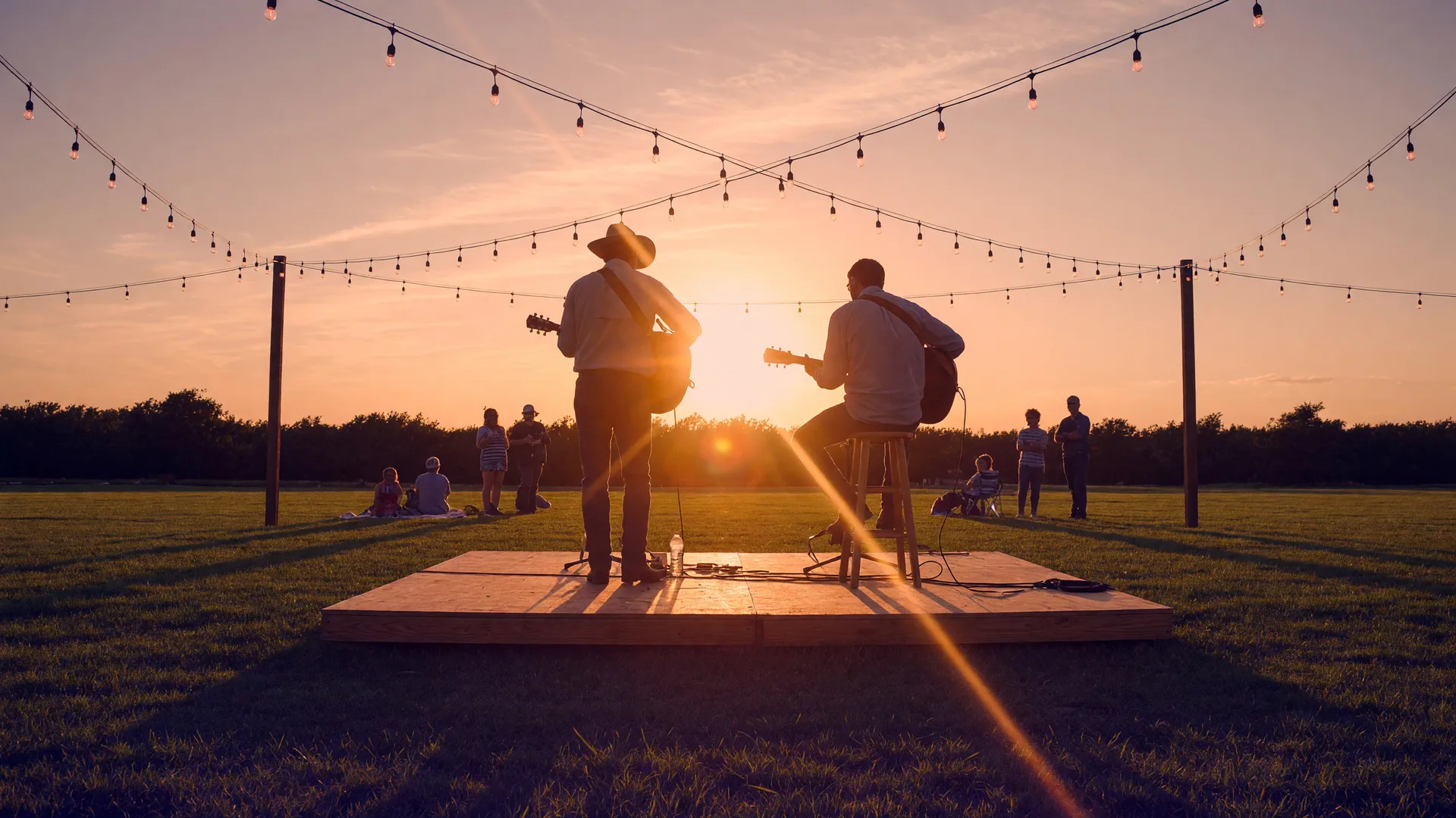 Two acoustic musicians silhouetted on an outdoor wooden stage at golden hour with string lights overhead