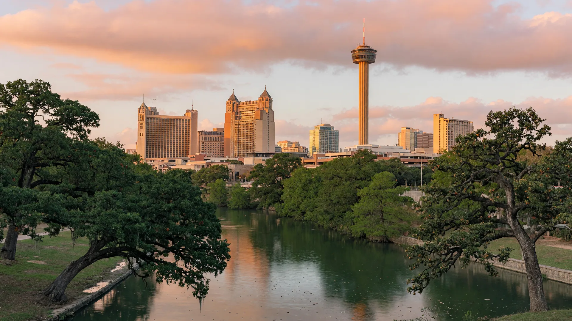 The downtown San Antonio skyline with the Tower of the Americas at golden hour, seen across a calm stretch of the San Antonio River and framed by live oaks