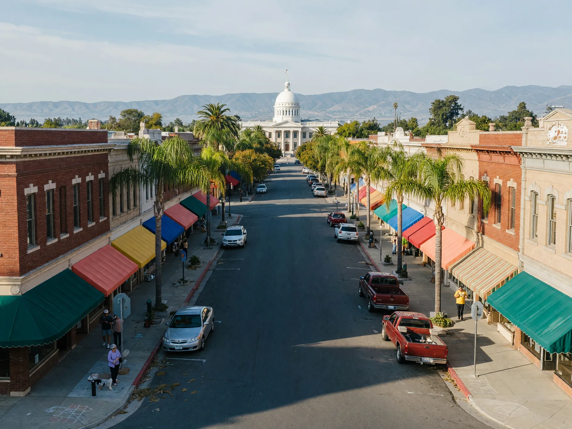 Main Street in downtown Merced on a bright late-summer morning with historic storefronts, palm-lined sidewalks, and the white dome of the Merced County Courthouse in the distance