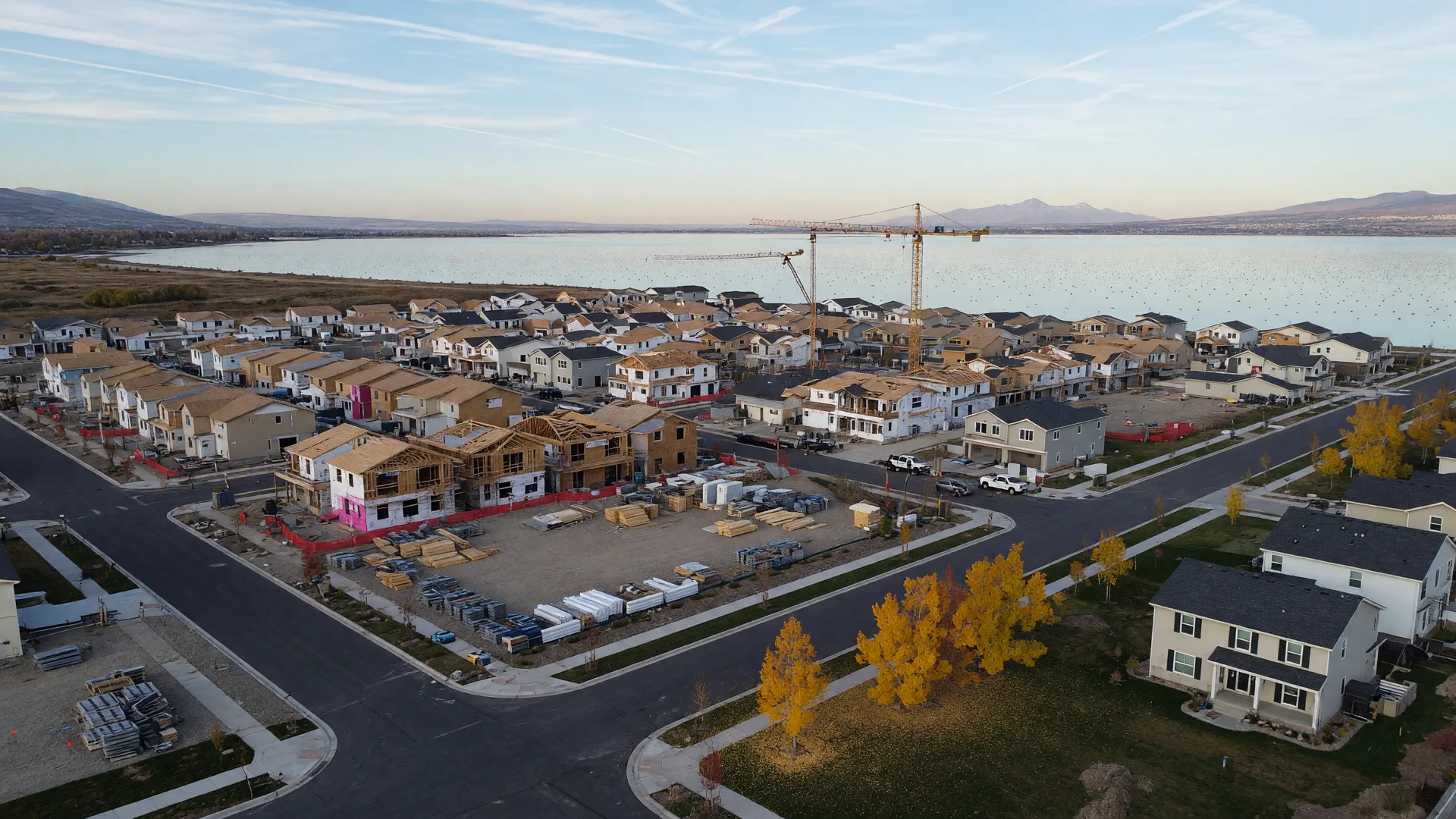 Aerial view of an active new-construction master-planned community in Utah County in late October with homes in framing and finishing stages, cranes, and Utah Lake in the distance