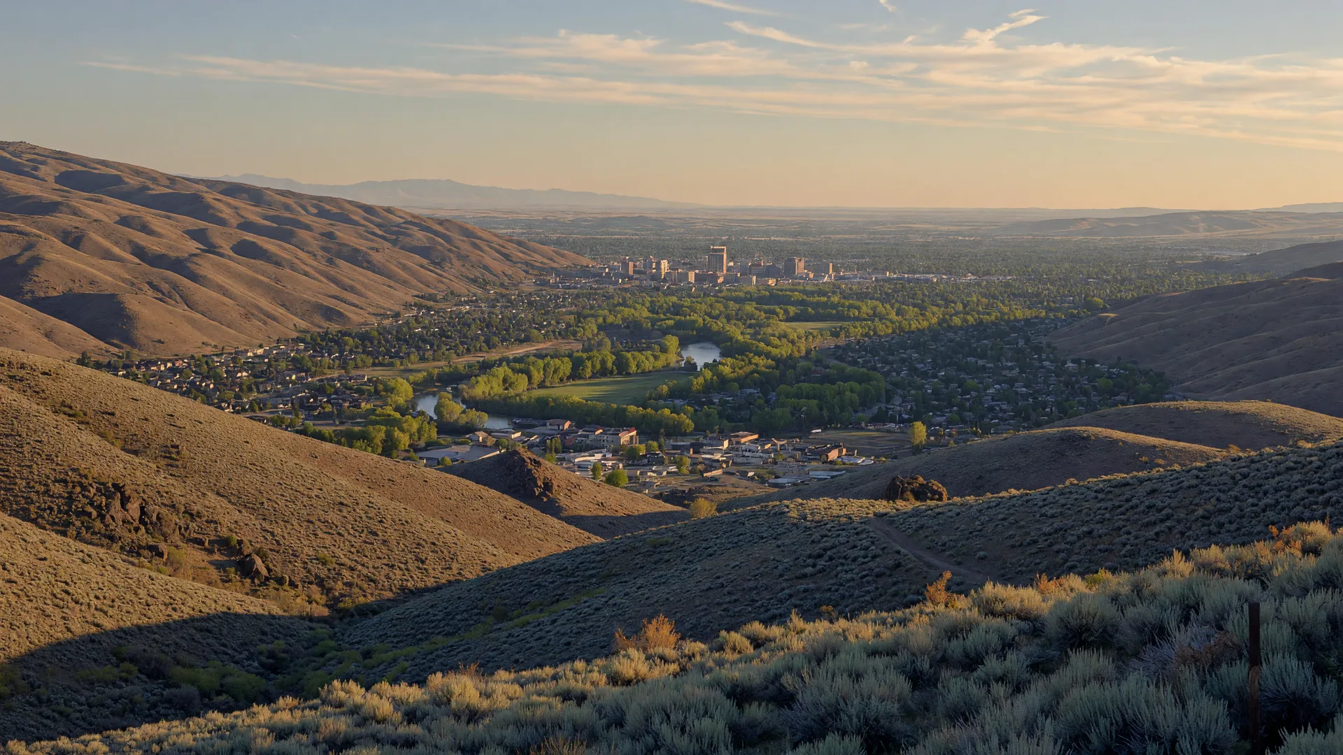Panoramic golden-hour valley view with rolling foothills, a winding river, and homes below a distant skyline