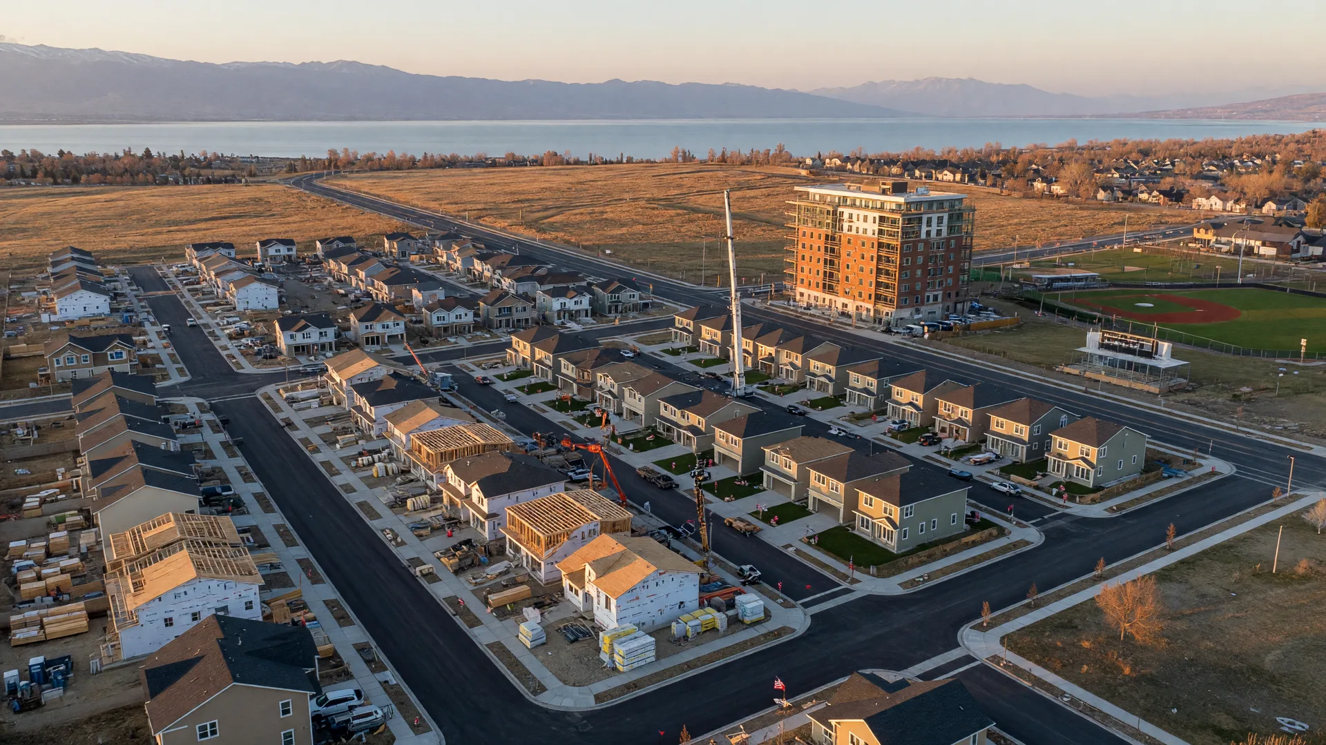 Aerial view of an active new-construction master plan along the Wasatch Front with homes in framing and finishing stages, fresh streets, and Utah Lake haze on the horizon